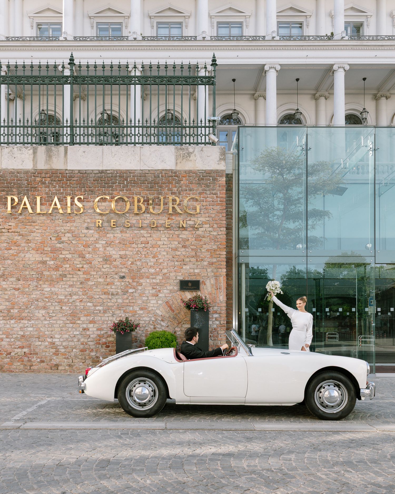 Bride in white gown poses in classic white convertible outside Palais Coburg Residenz in Vienna.
