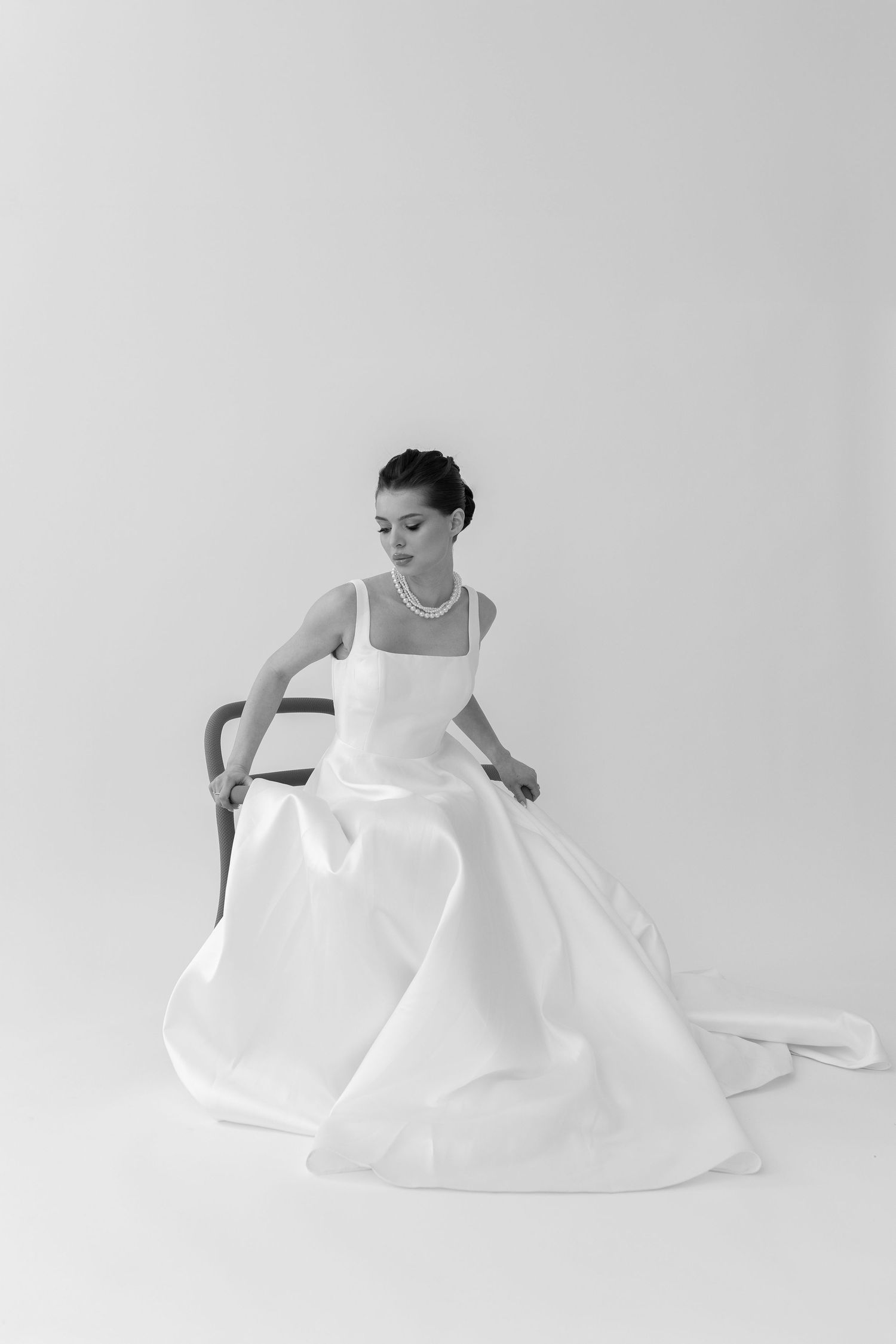 Elegant bride in a minimalist white ball gown with pearl necklace seated on a chair in a black and white studio photo.