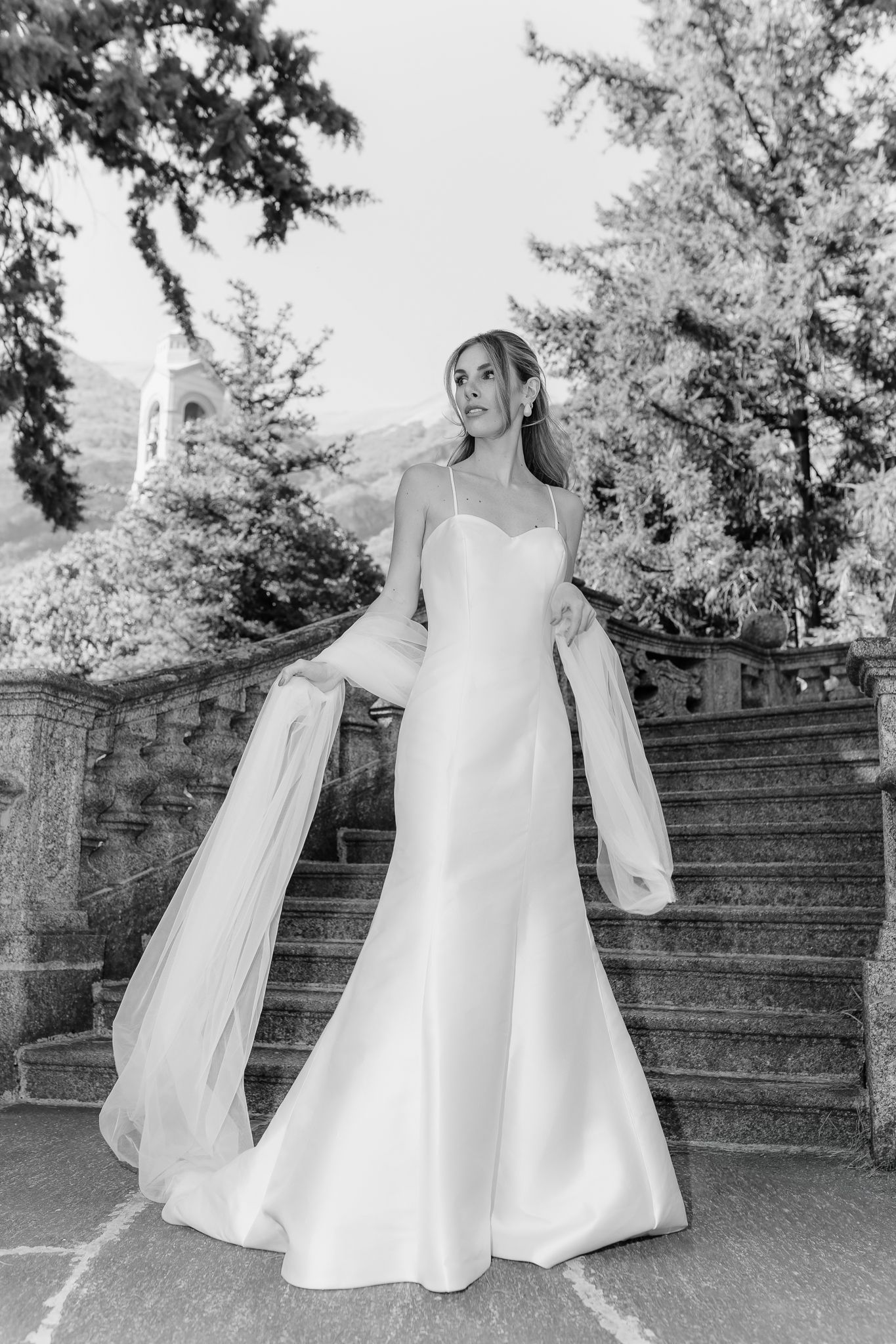 Black and white portrait of bride in mermaid gown with flowing sheer wrap on ornate stone stairs.