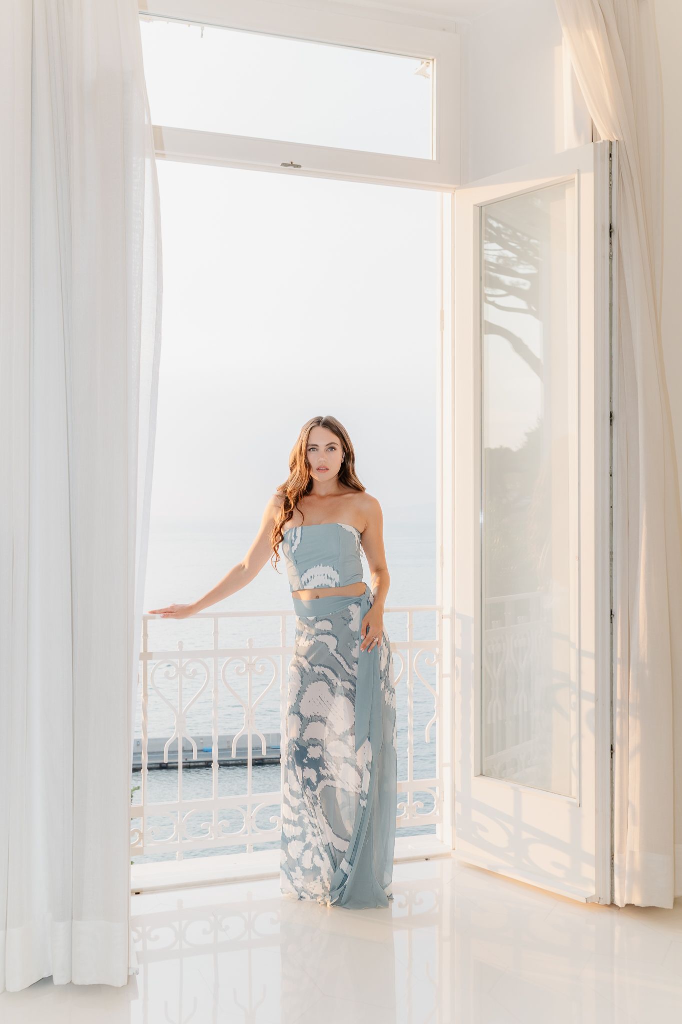 Woman in blue floral two-piece outfit standing in open white French doors with ocean view balcony.