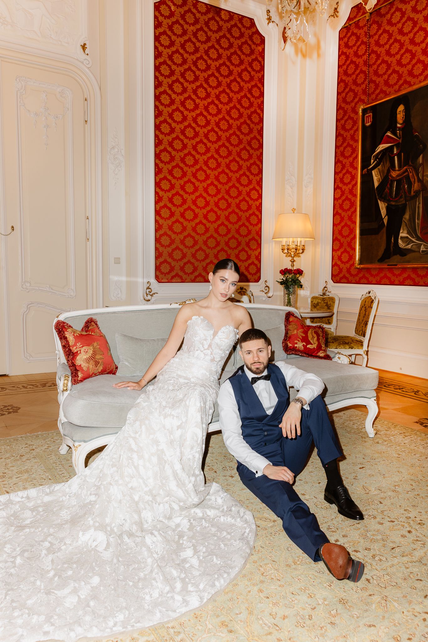 Bride in lace gown and groom in navy suit pose together on ornate sofa in red damask luxury room of Hotel Imperial in Vienna.