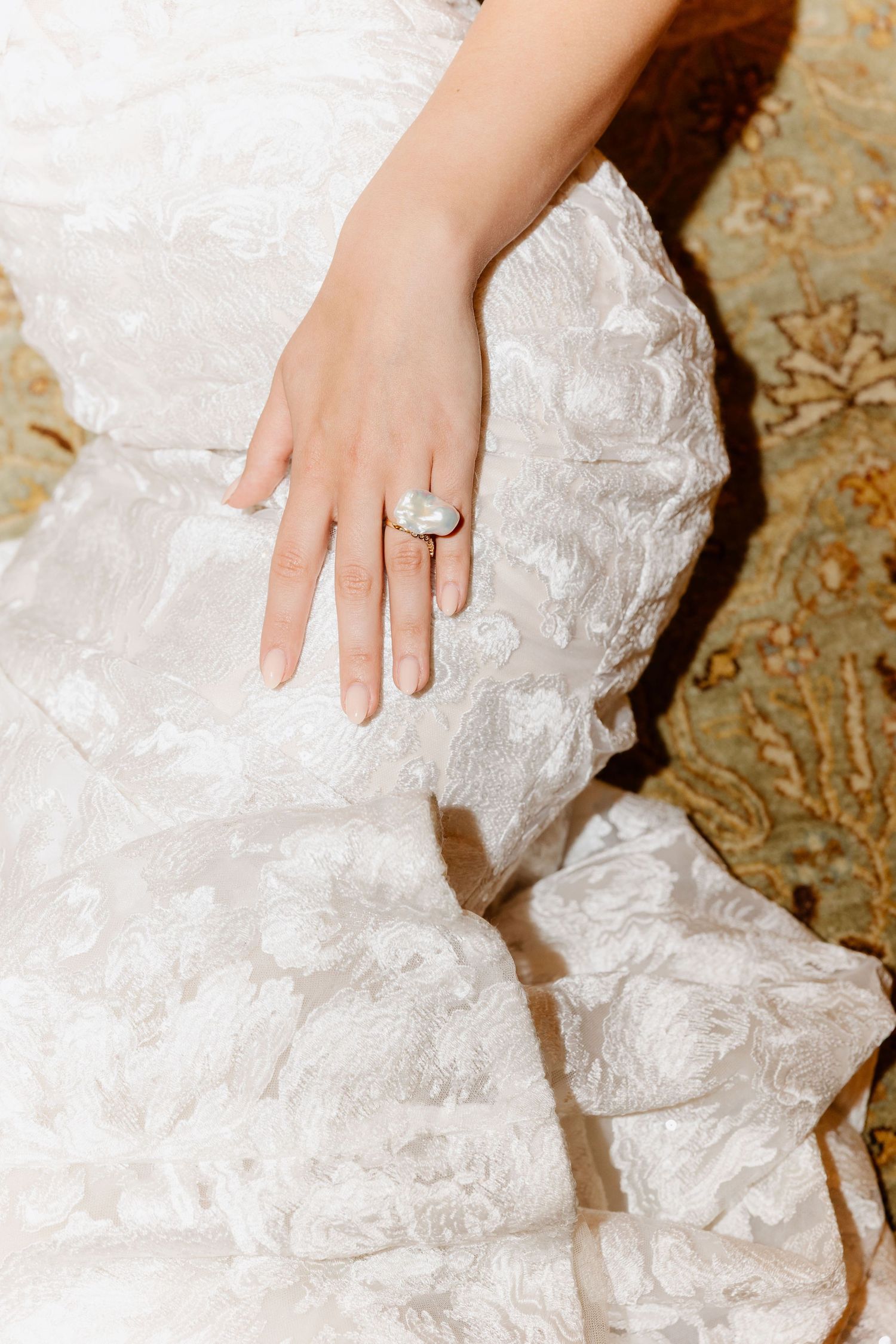 Close-up of bride's hand wearing a large oval moonstone ring resting on her lace wedding gown.