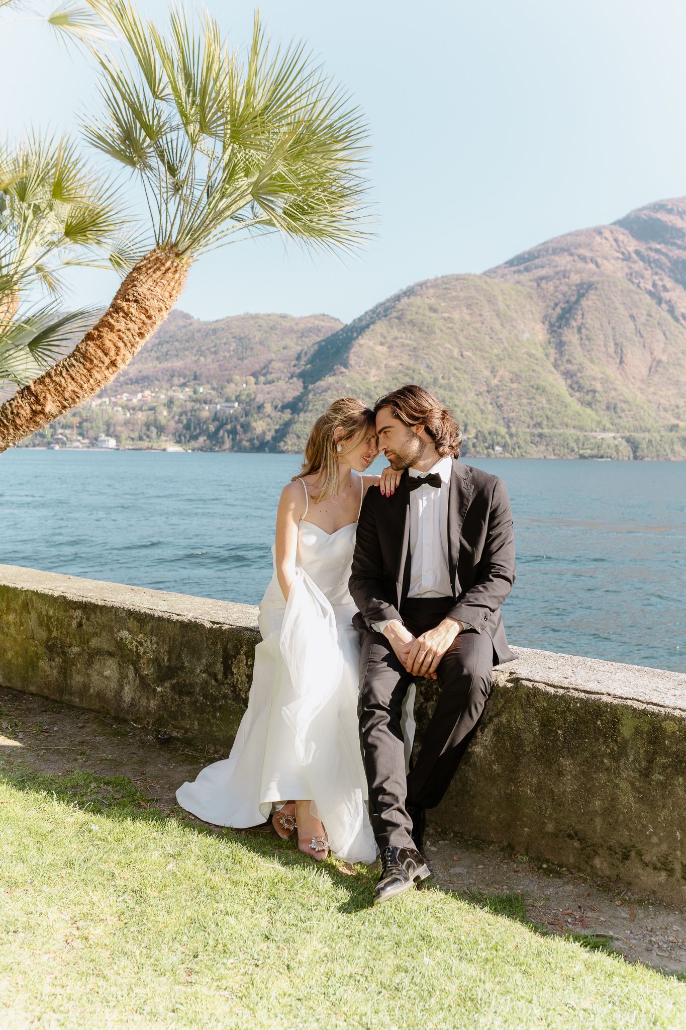 Bride and groom share an intimate forehead touch seated on a lakeside wall with palm tree and mountains.