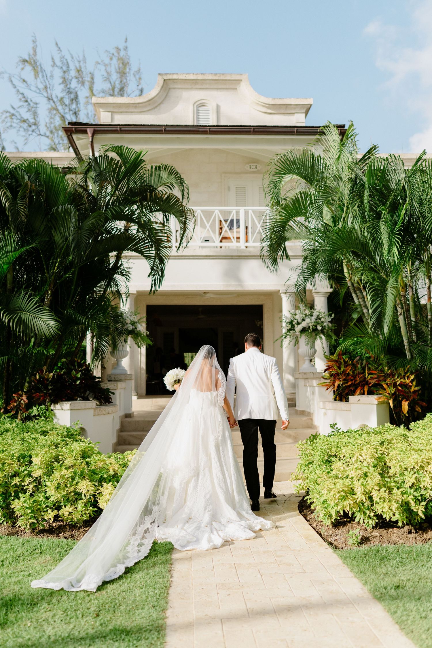 Bride in cathedral veil and groom in white jacket walk toward tropical colonial wedding venue. Barbados Destination Wedding Photographer