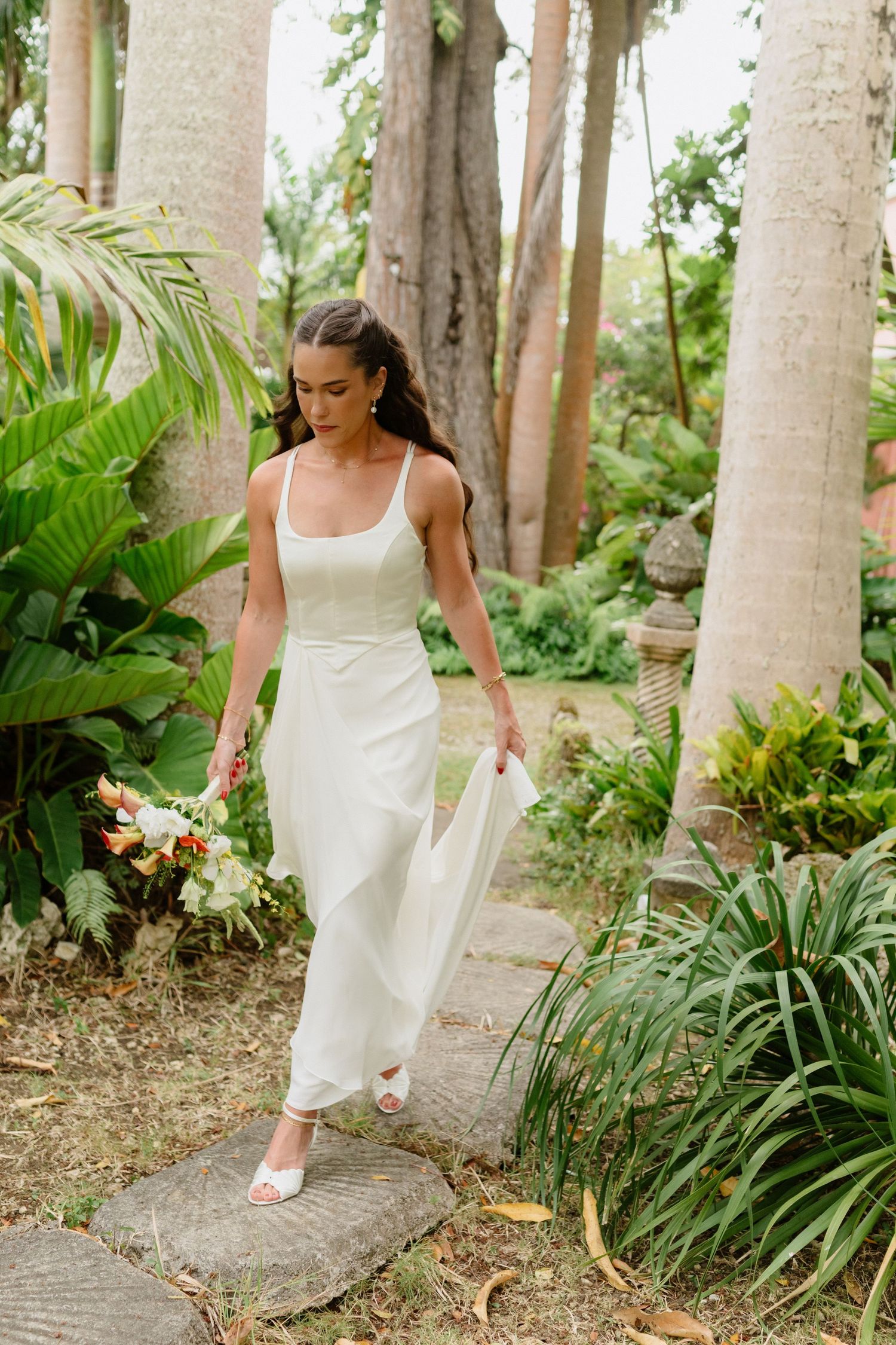 Bride in elegant white slip dress walks through lush tropical garden holding colorful bouquet. Barbados photographer