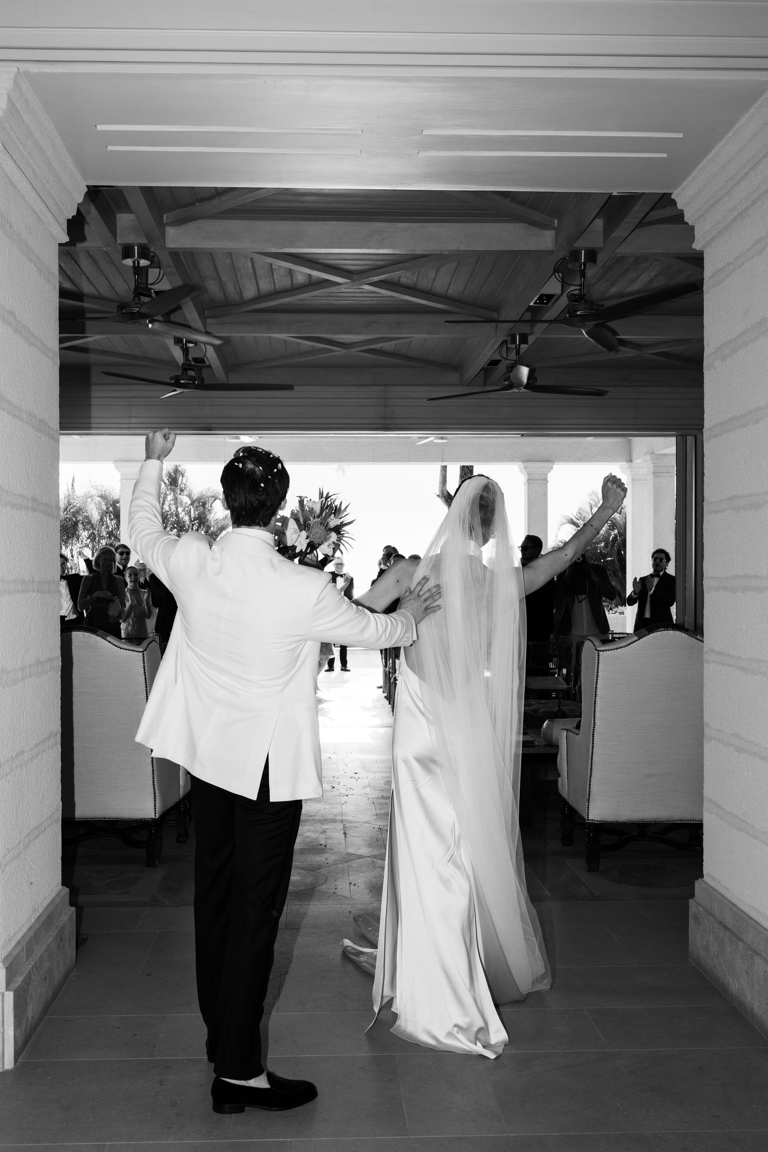 Black and white photo of bride and groom raising arms in celebration as they exit wedding ceremony. Barbados Destination Wedding Photographe