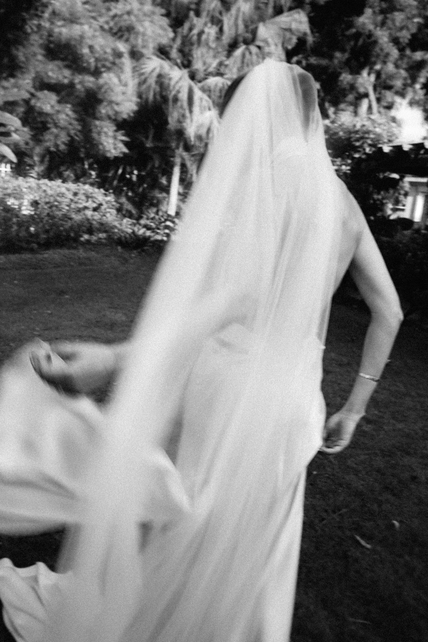 Bride in flowing white gown and veil walking through lush tropical garden in artistic black and white. Barbados photographer