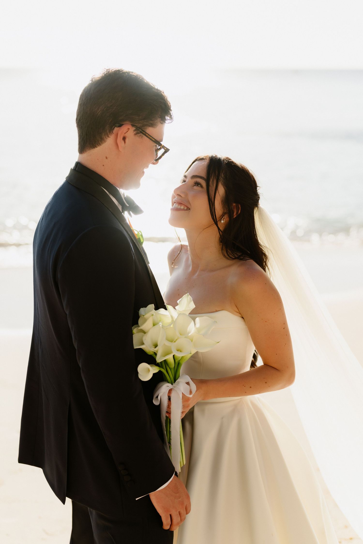 Bride holding white calla lily bouquet gazes lovingly at groom on sunlit beach after ceremony. Barbados Destination Wedding Photographer