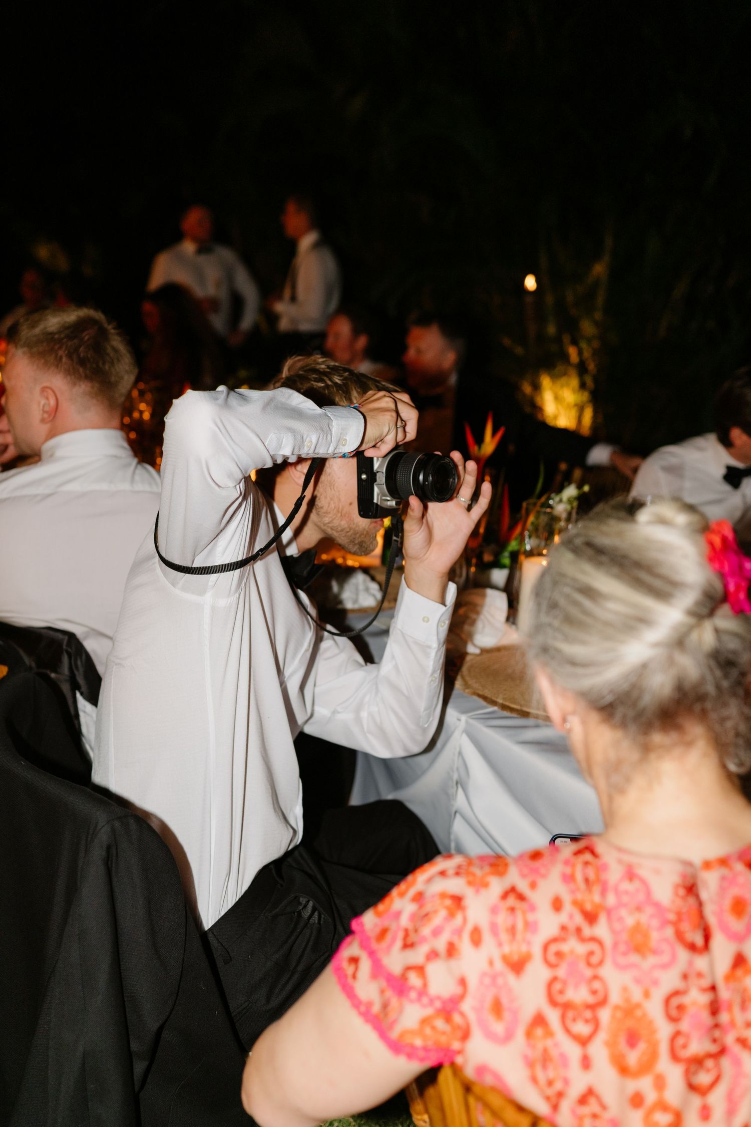Photographer capturing shots at an evening wedding reception with guests seated at decorated tables in a dimly lit outdoor venue. Barbados