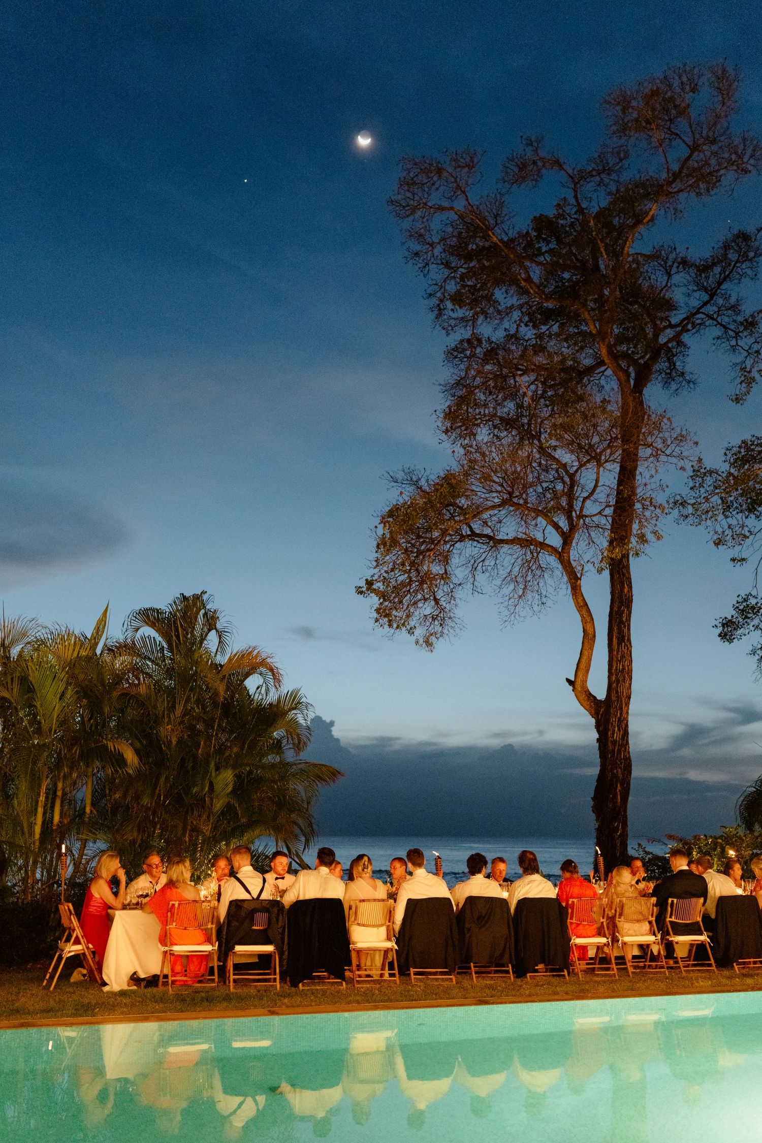 Outdoor wedding reception dinner by a pool at twilight with guests seated under a moonlit sky. Barbados photographer