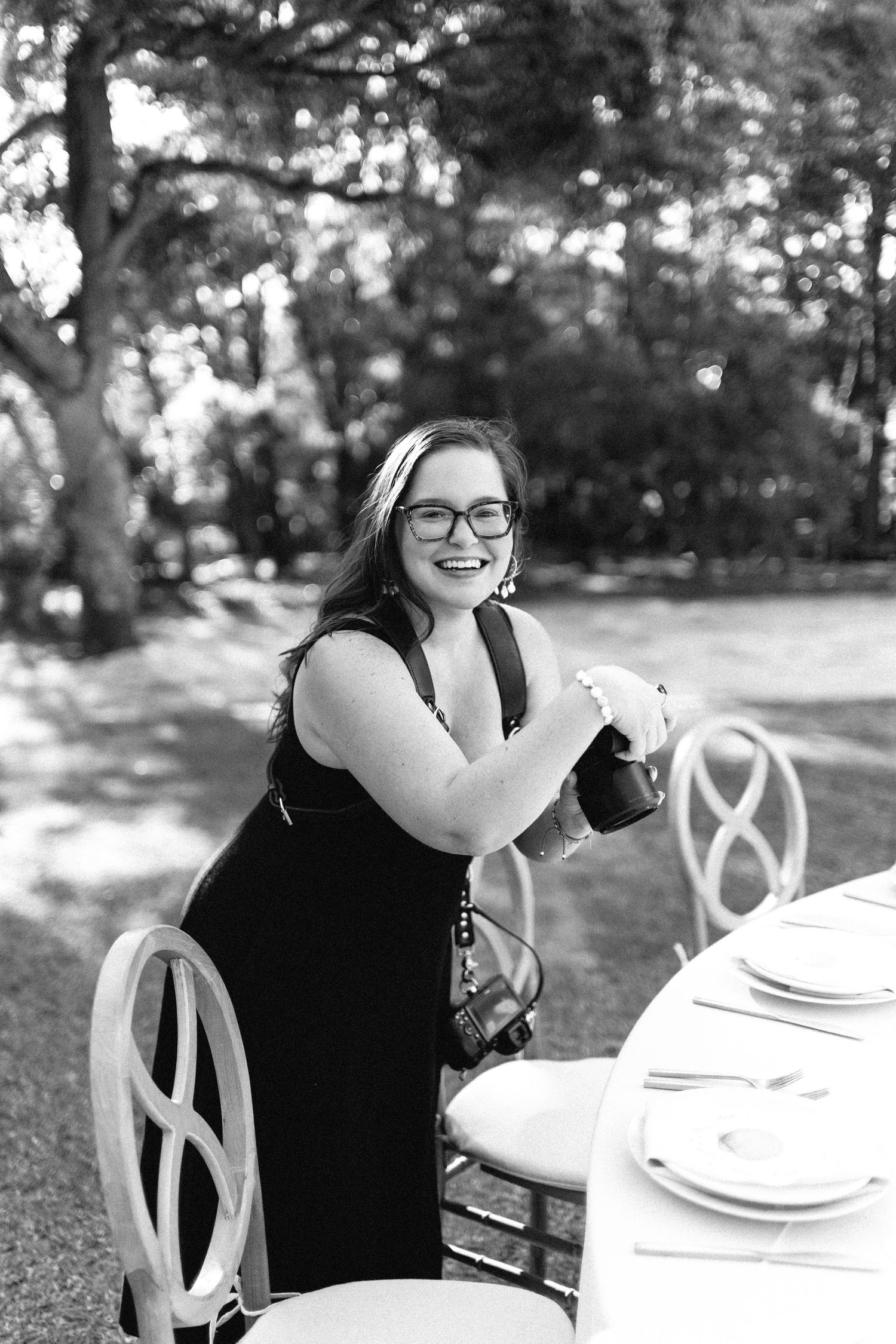 Black and white photo of smiling woman with glasses holding camera at outdoor wedding reception. Barbados Destination Wedding Photographer