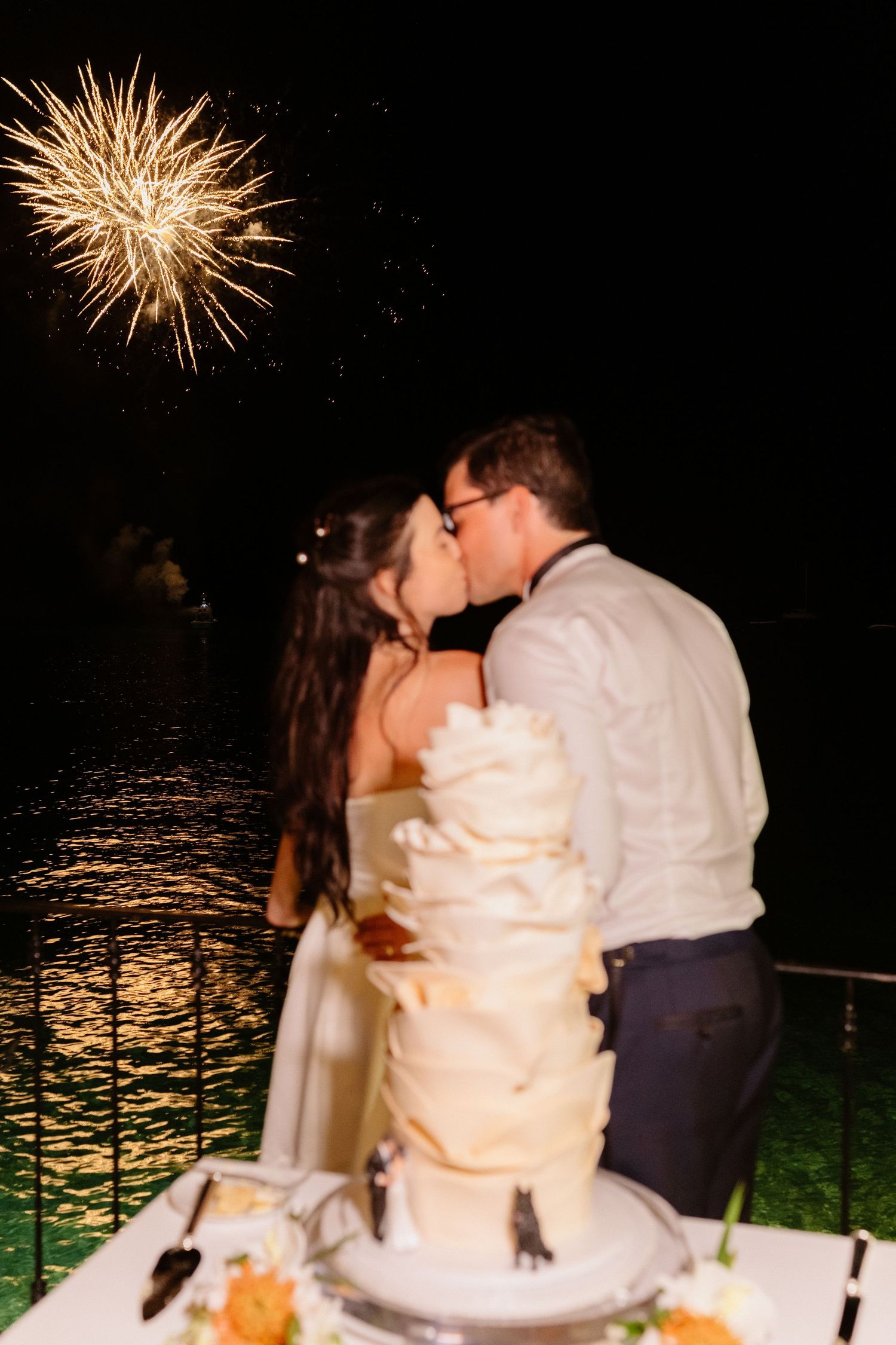 Newlyweds kiss beside tiered wedding cake with fireworks exploding over water in night sky. Barbados Destination Wedding Photographer