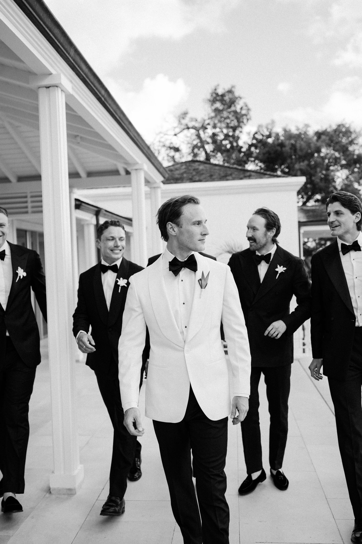 Groom in white tuxedo jacket leads groomsmen in black tuxedos walking outdoors at elegant wedding venue. Barbados Wedding Photographer