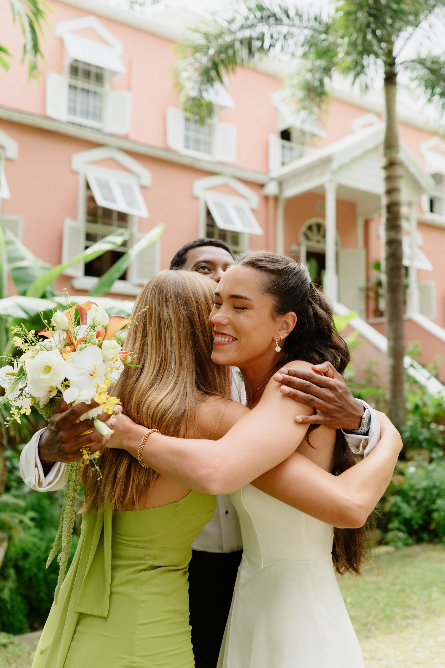 Bride and guest embrace warmly outside pink colonial building during tropical wedding celebration. Barbados Destination Wedding Photographer