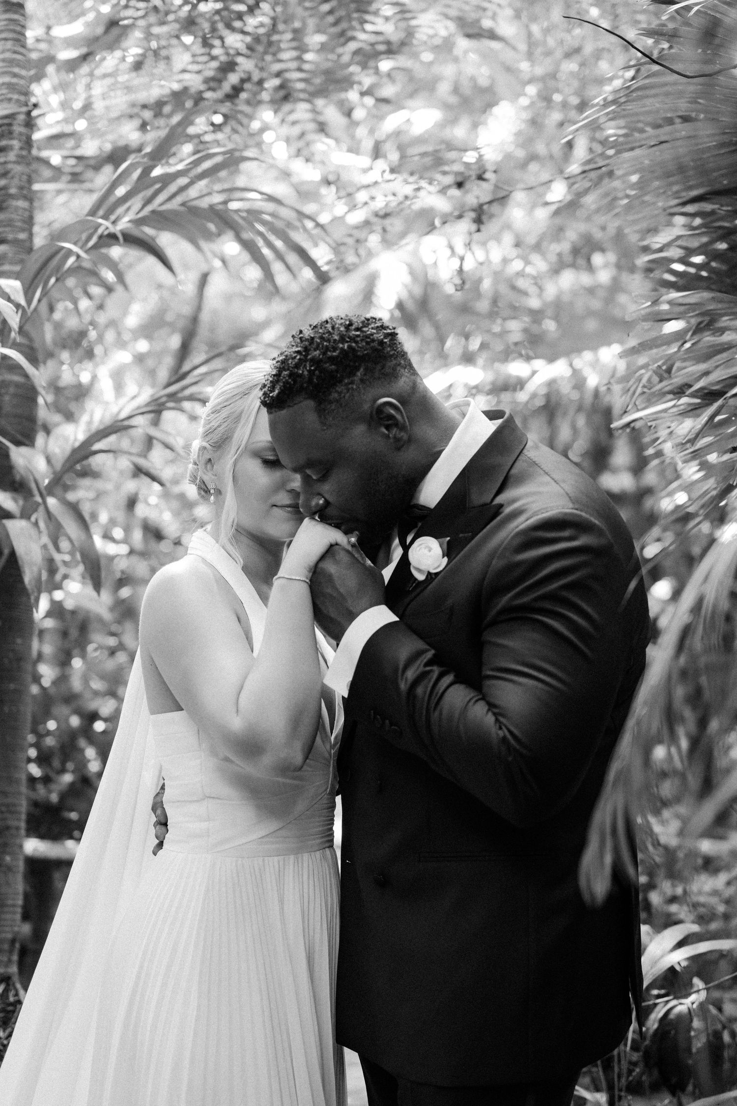 Black and white photo of groom tenderly kissing bride's hand in a lush tropical garden setting. Barbados photographer