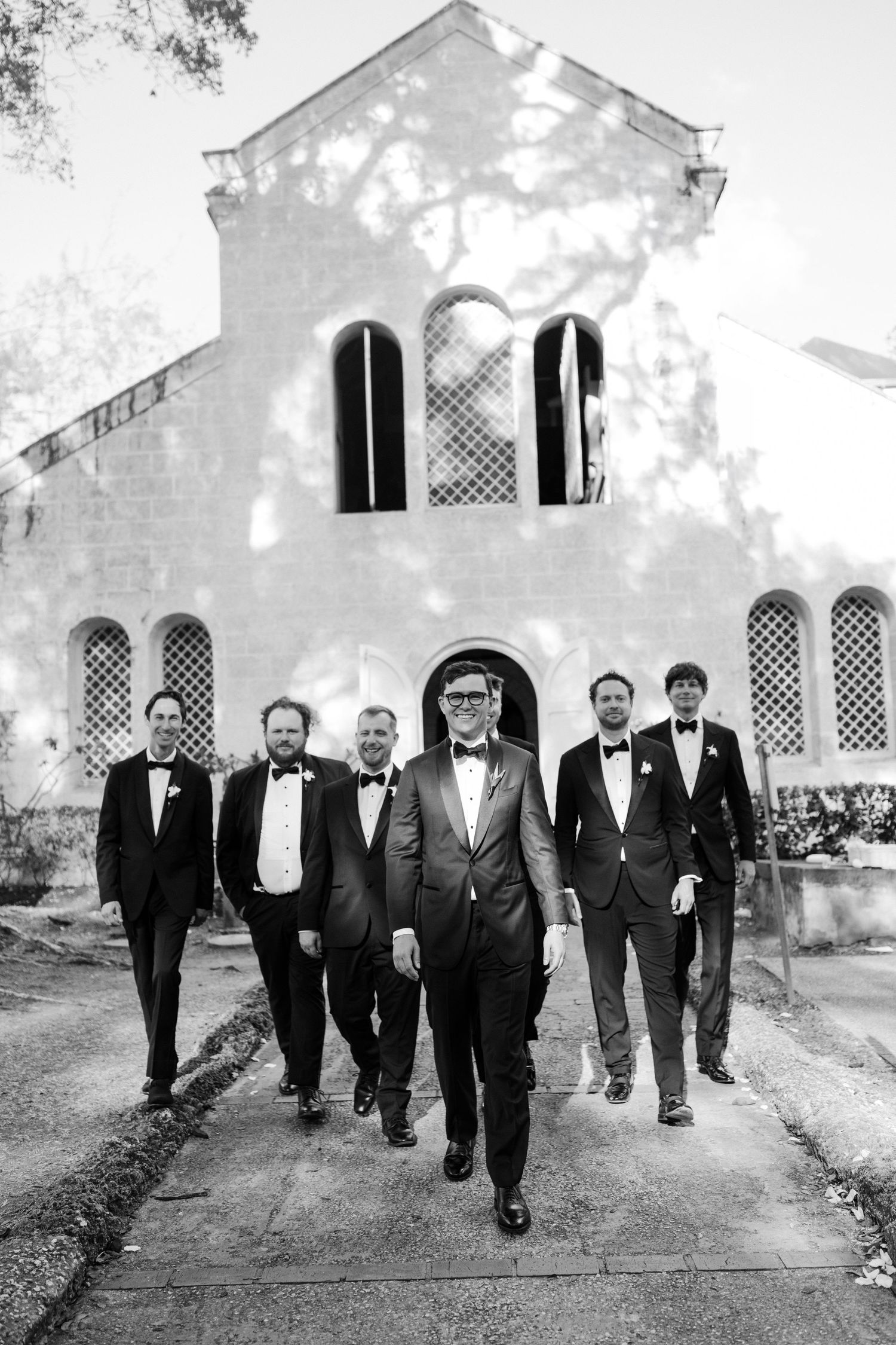Groom leads groomsmen in tuxedos walking confidently before a historic church in black and white. Barbados Destination Wedding Photographer