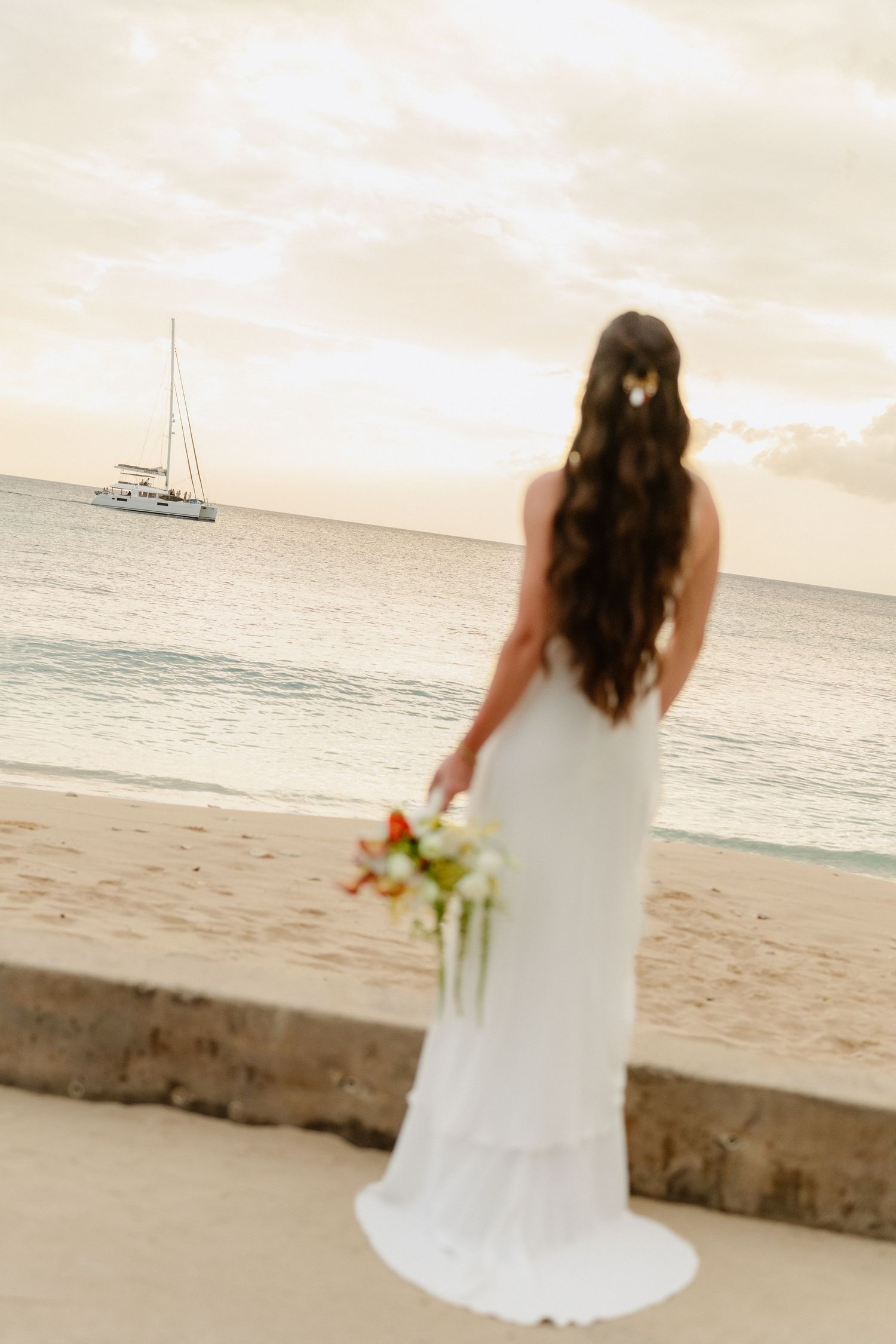 Elegant bride in white gown holding bouquet gazing at sailboat on calm ocean at sunset. Barbados Destination Wedding Photographer