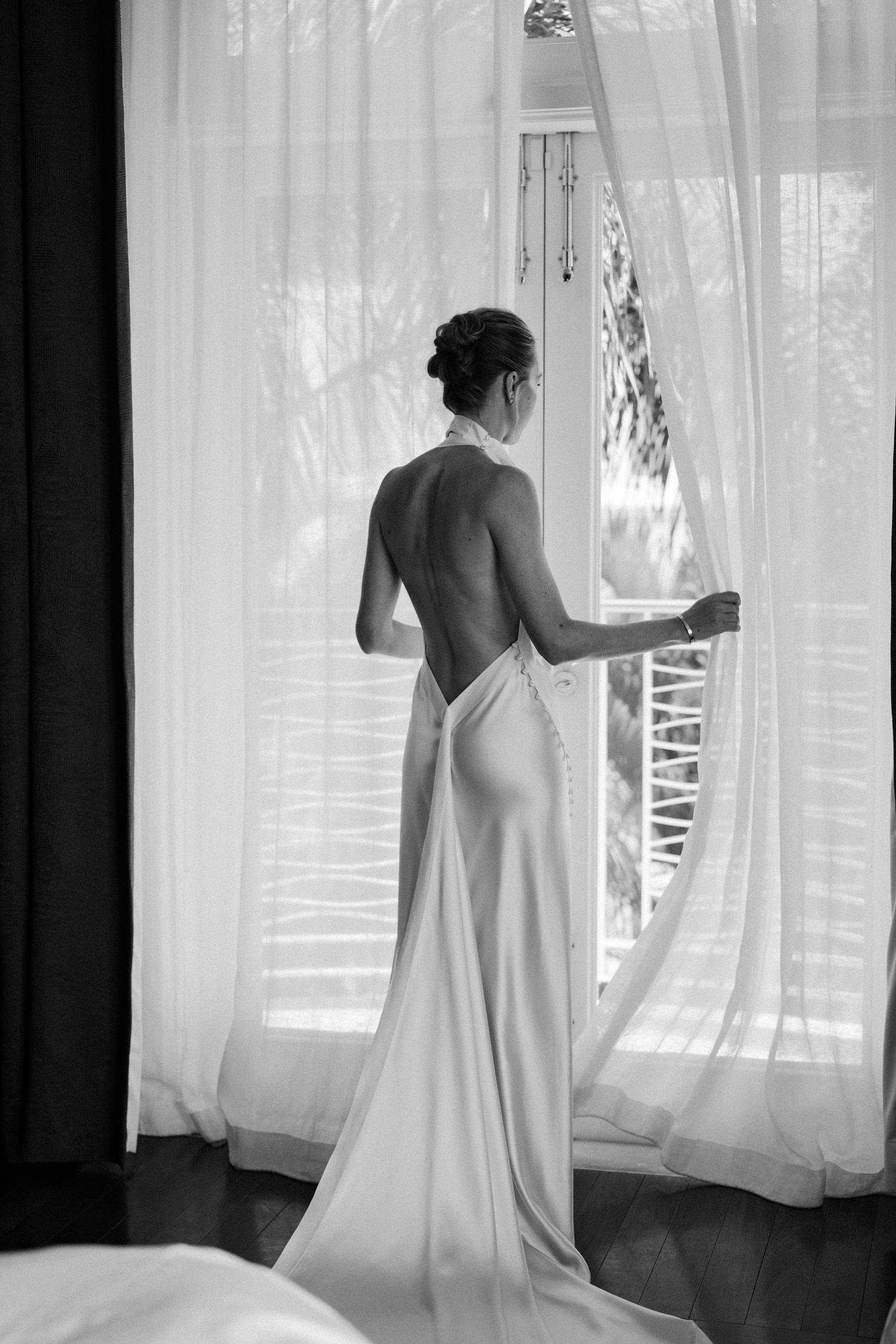 Black and white photo of bride in backless halter wedding gown standing at a sunlit window. Barbados Destination Wedding Photographer