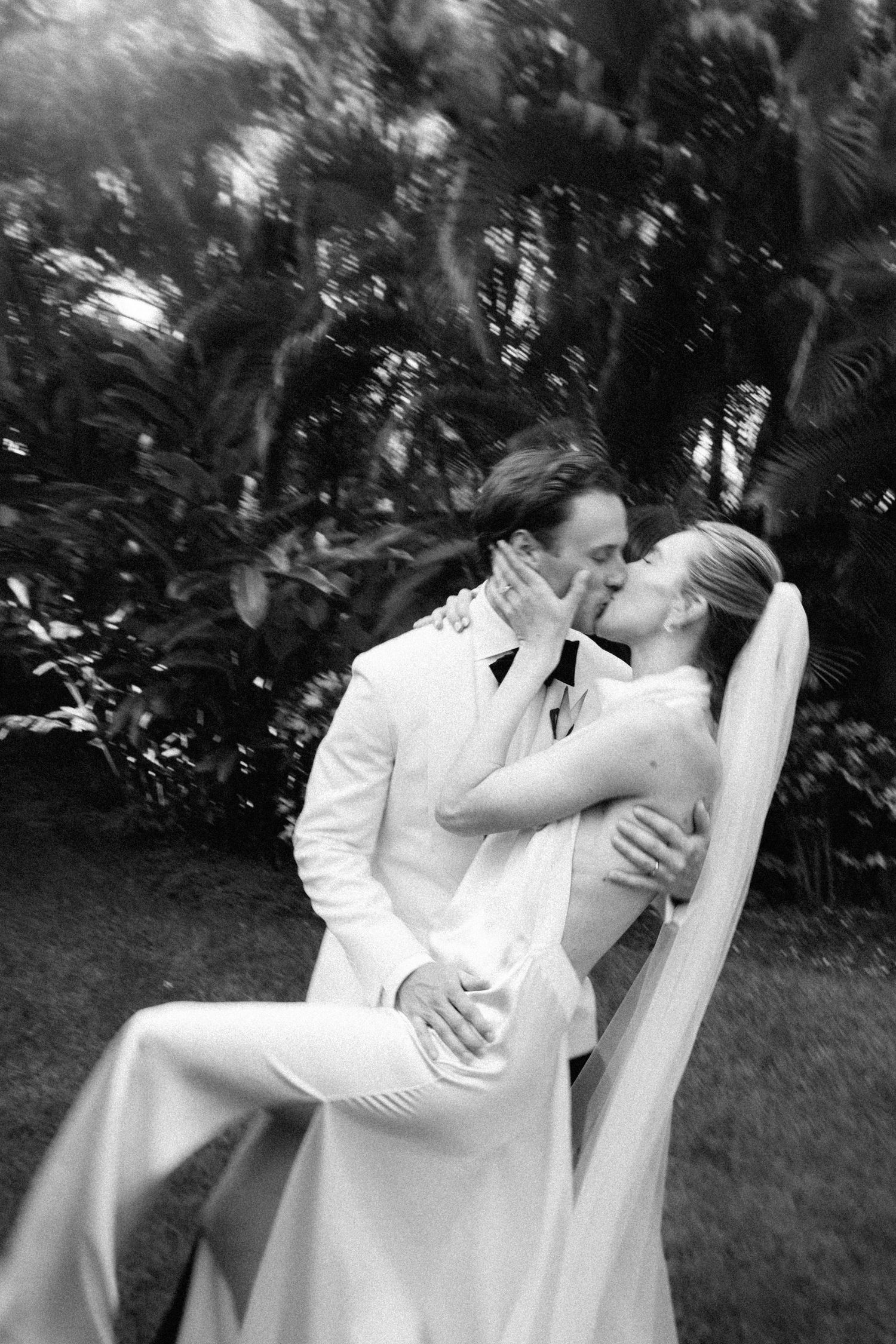 Bride and groom share a romantic kiss outdoors, with lush tropical greenery in this elegant black and white wedding photo. Barbados