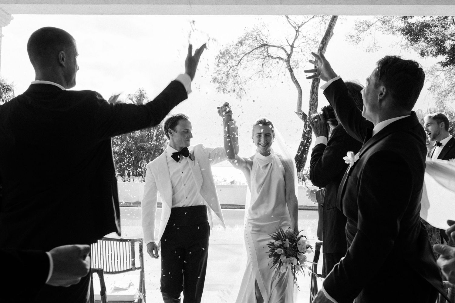 Newlyweds celebrate with confetti as groomsmen cheer in black and white wedding ceremony photo. Barbados photographer