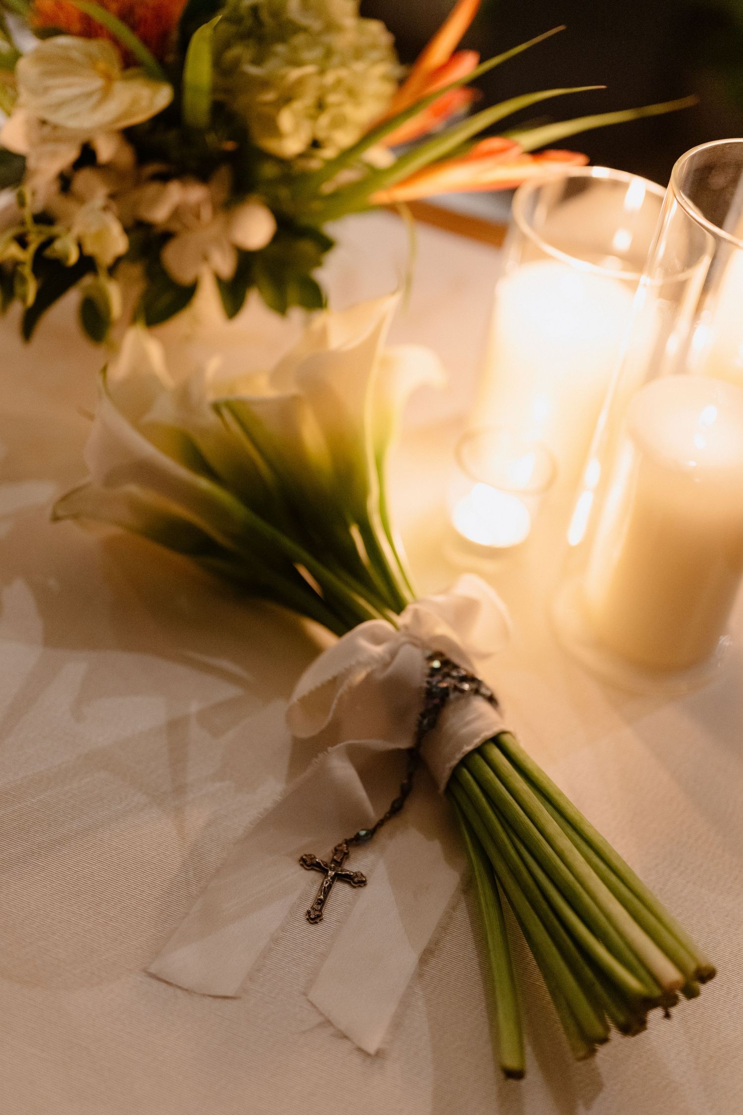 Bridal bouquet of white calla lilies with cross charm rests beside glowing candles on wedding table. Barbados Destination Wedding Photograph