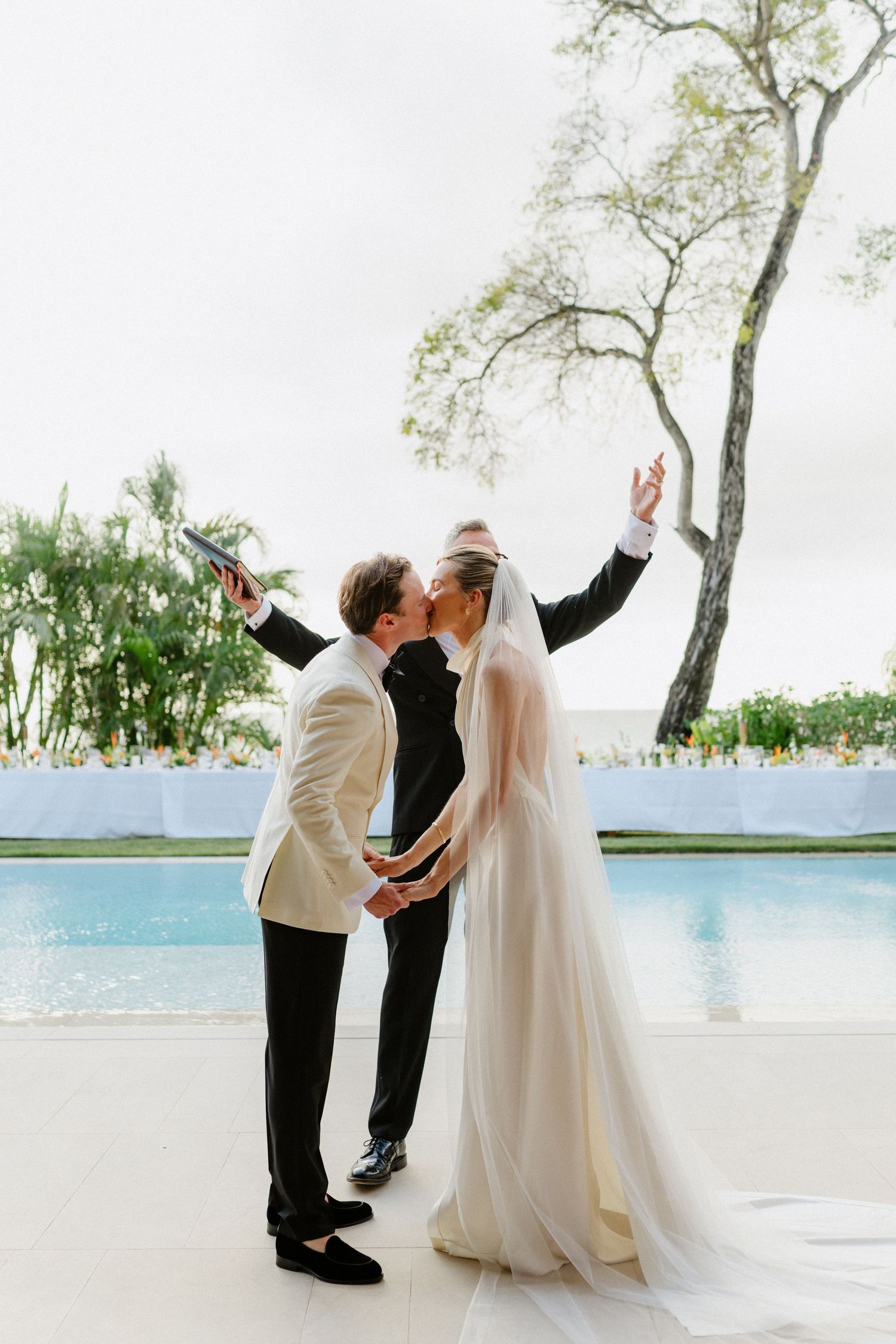 Groom in white jacket kisses bride in flowing veil poolside as officiant celebrates behind them. Barbados Destination Wedding Photographer