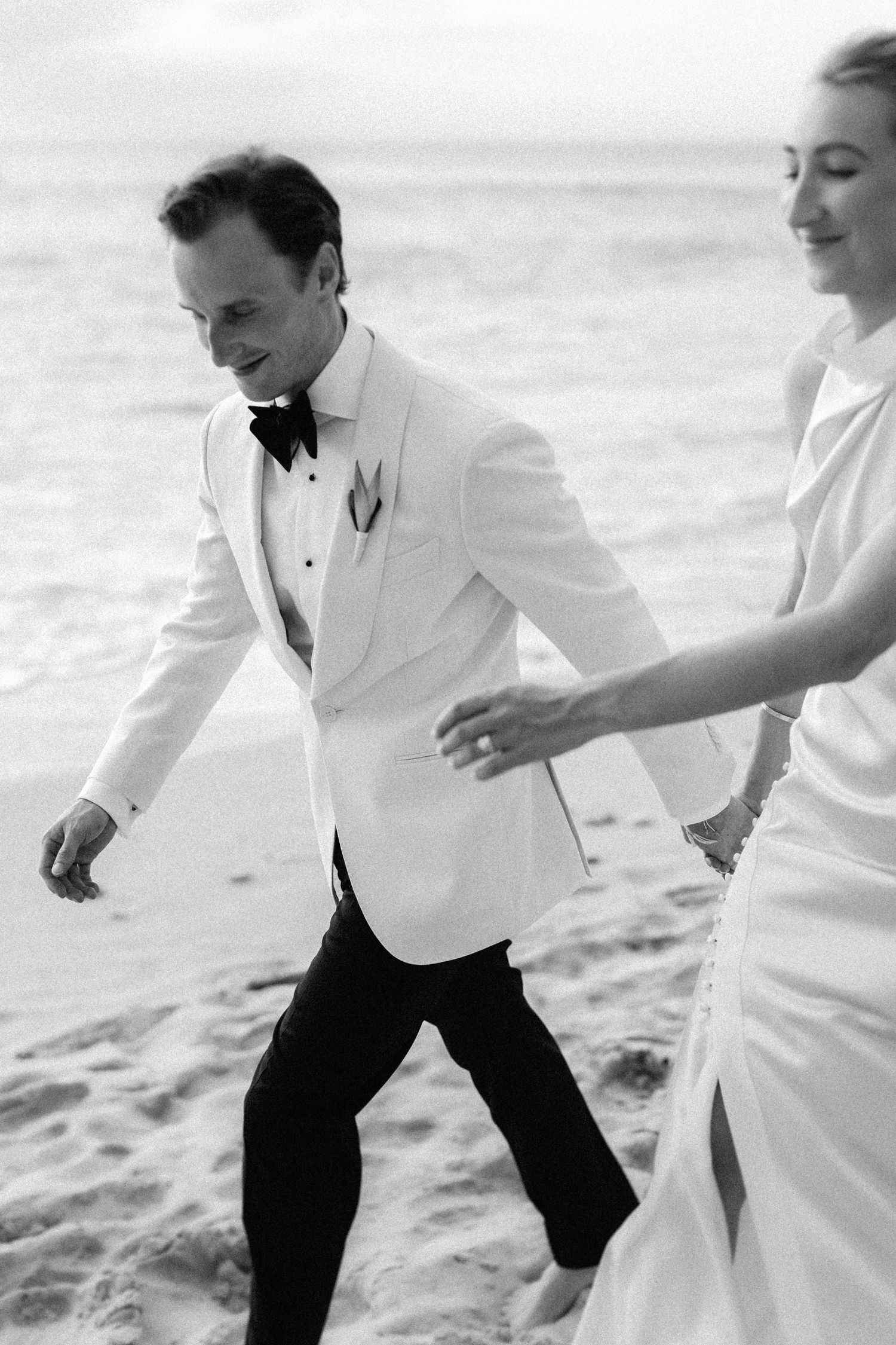 Bride and groom walking on beach in black and white, groom wearing white tuxedo jacket and black bow tie. Barbados photographer