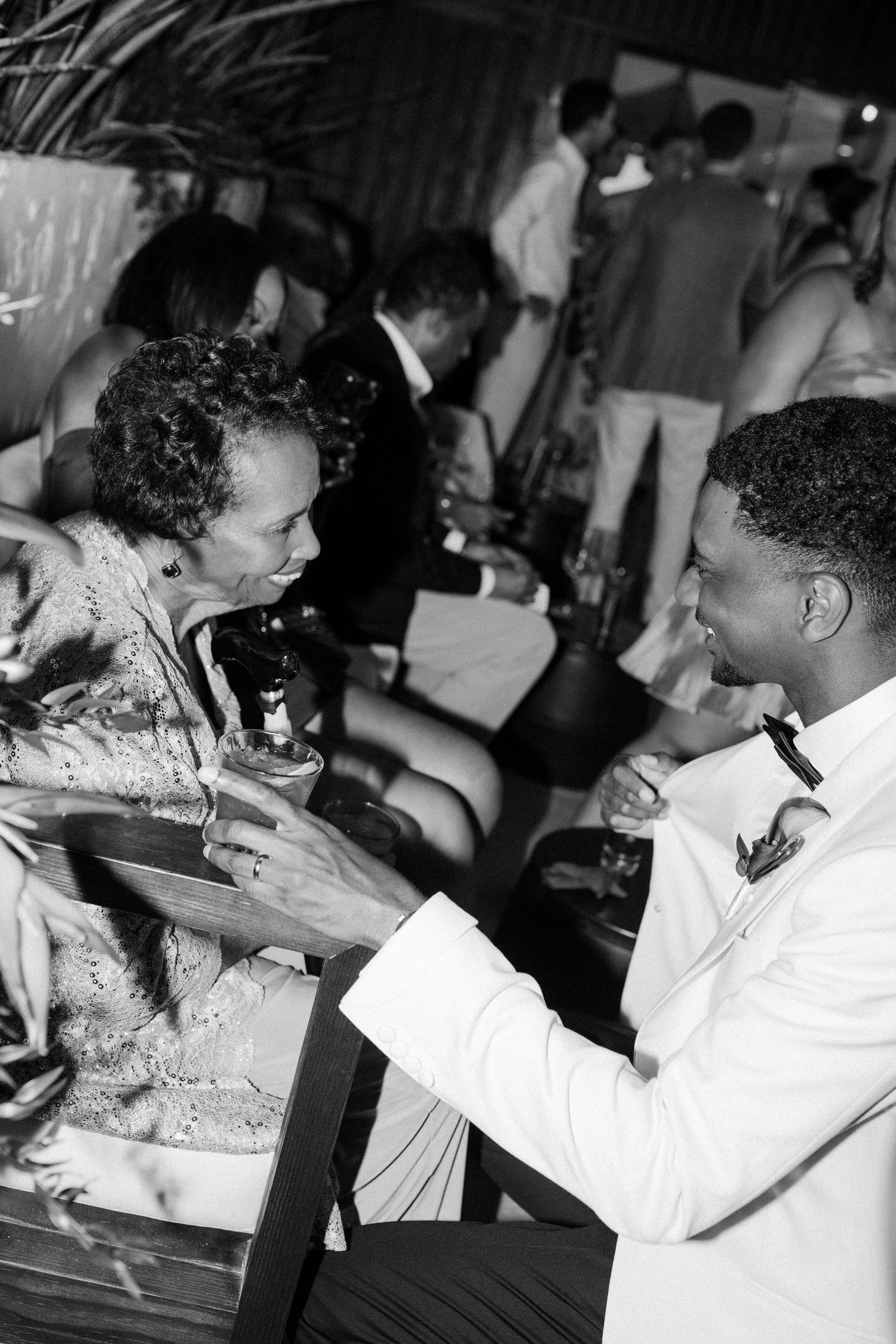 Black and white photo of groom in white tuxedo conversing with guest at wedding reception. Barbados photographer