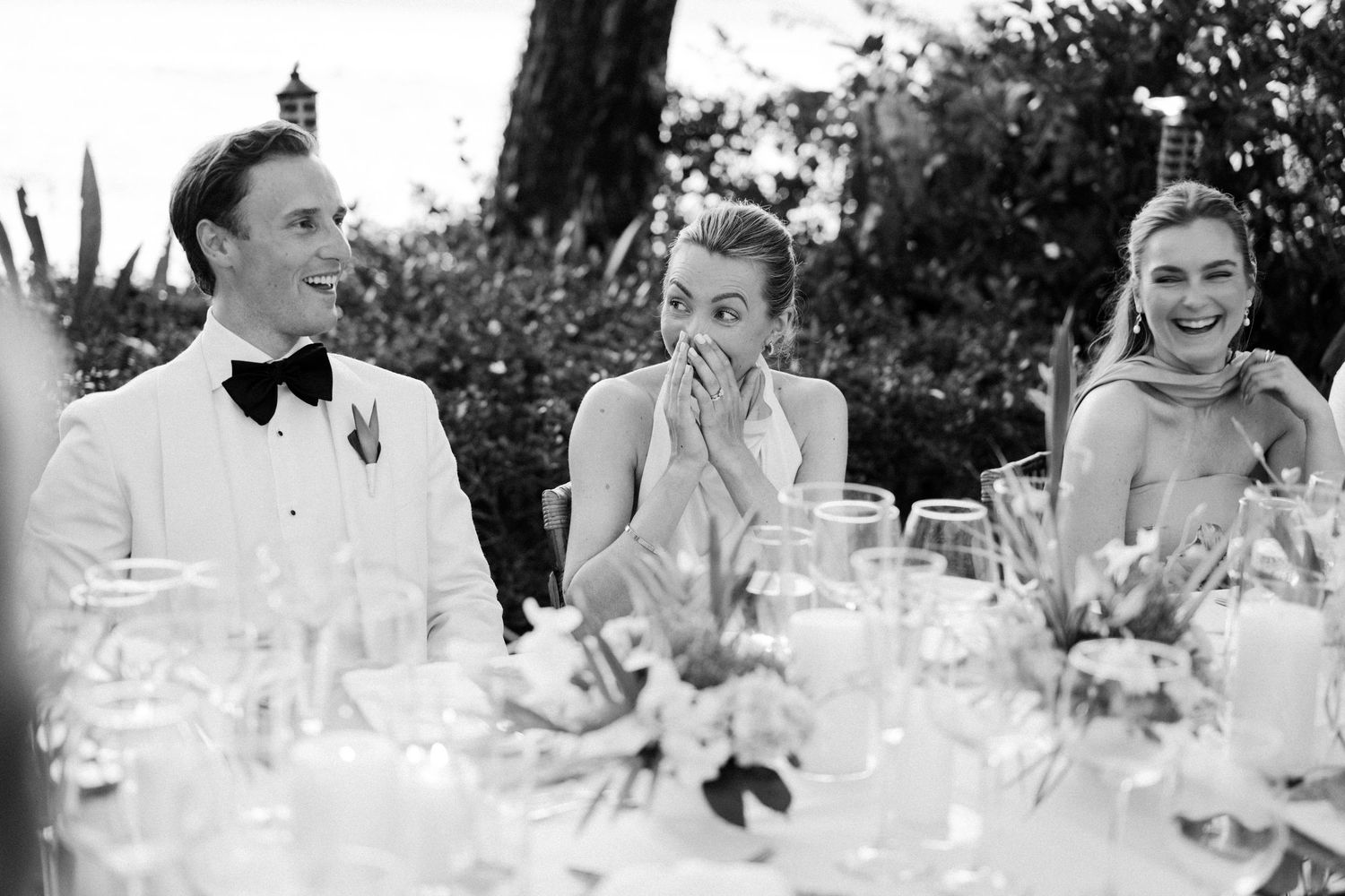 Black and white photo of bride and groom laughing with guests at an outdoor wedding reception dinner. Barbados photographer
