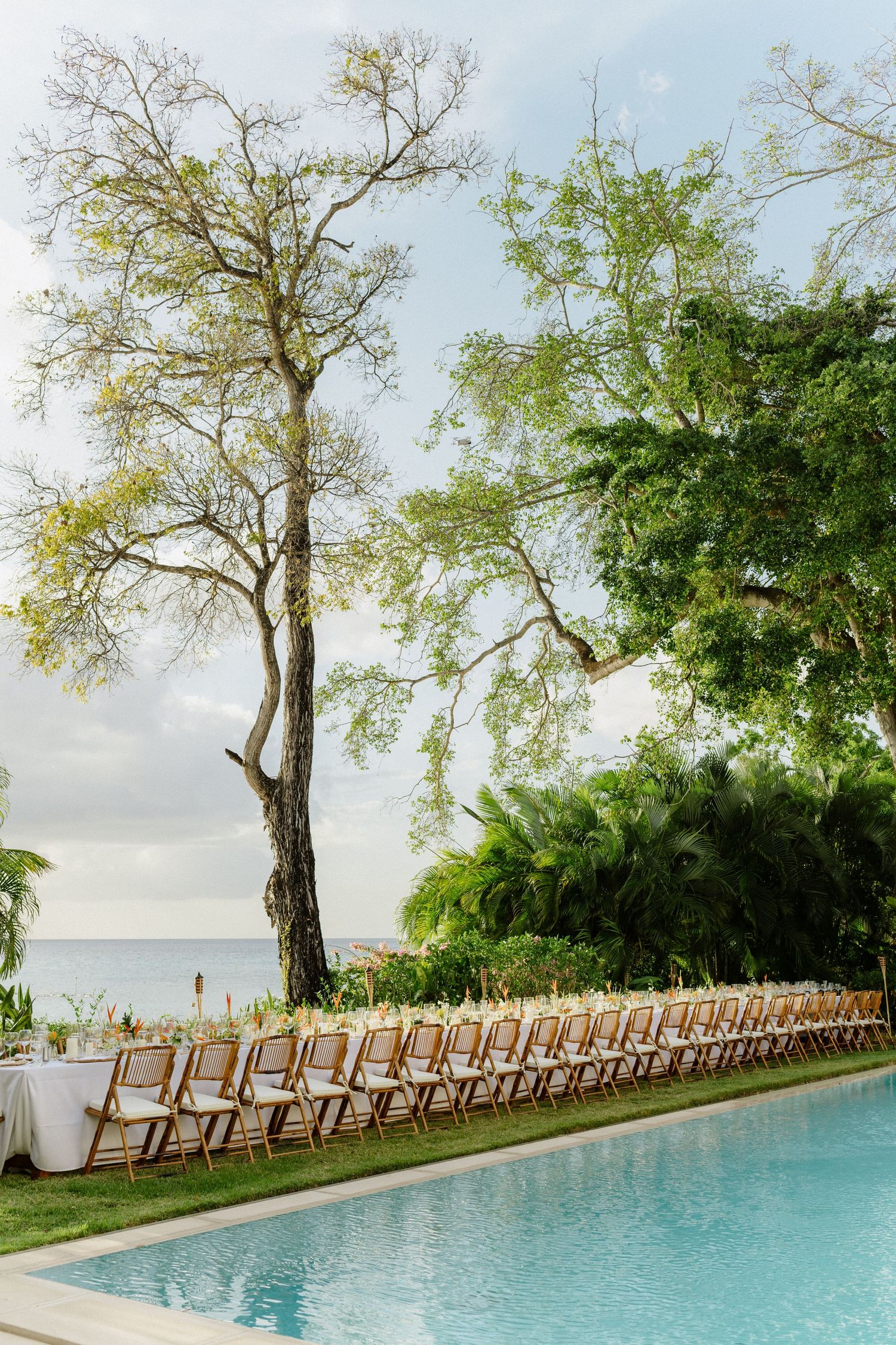 Long wedding reception table with wooden chairs beside a pool overlooking the ocean at sunset. Barbados Destination Wedding Photographer