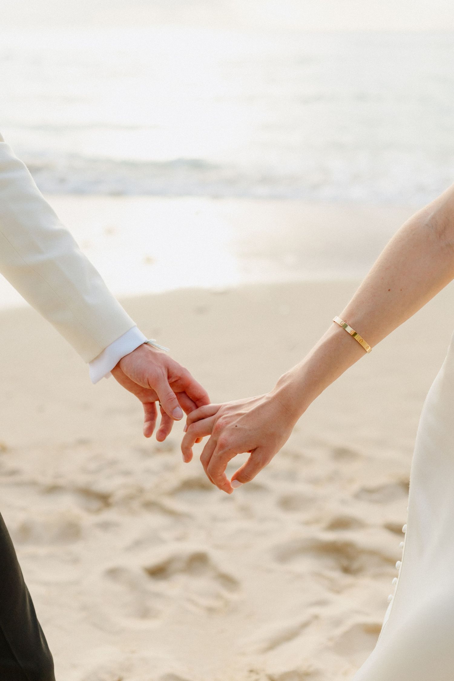 Close-up of bride and groom holding hands on sandy beach, bride wearing gold bracelet and wedding ring. West Coast barbados