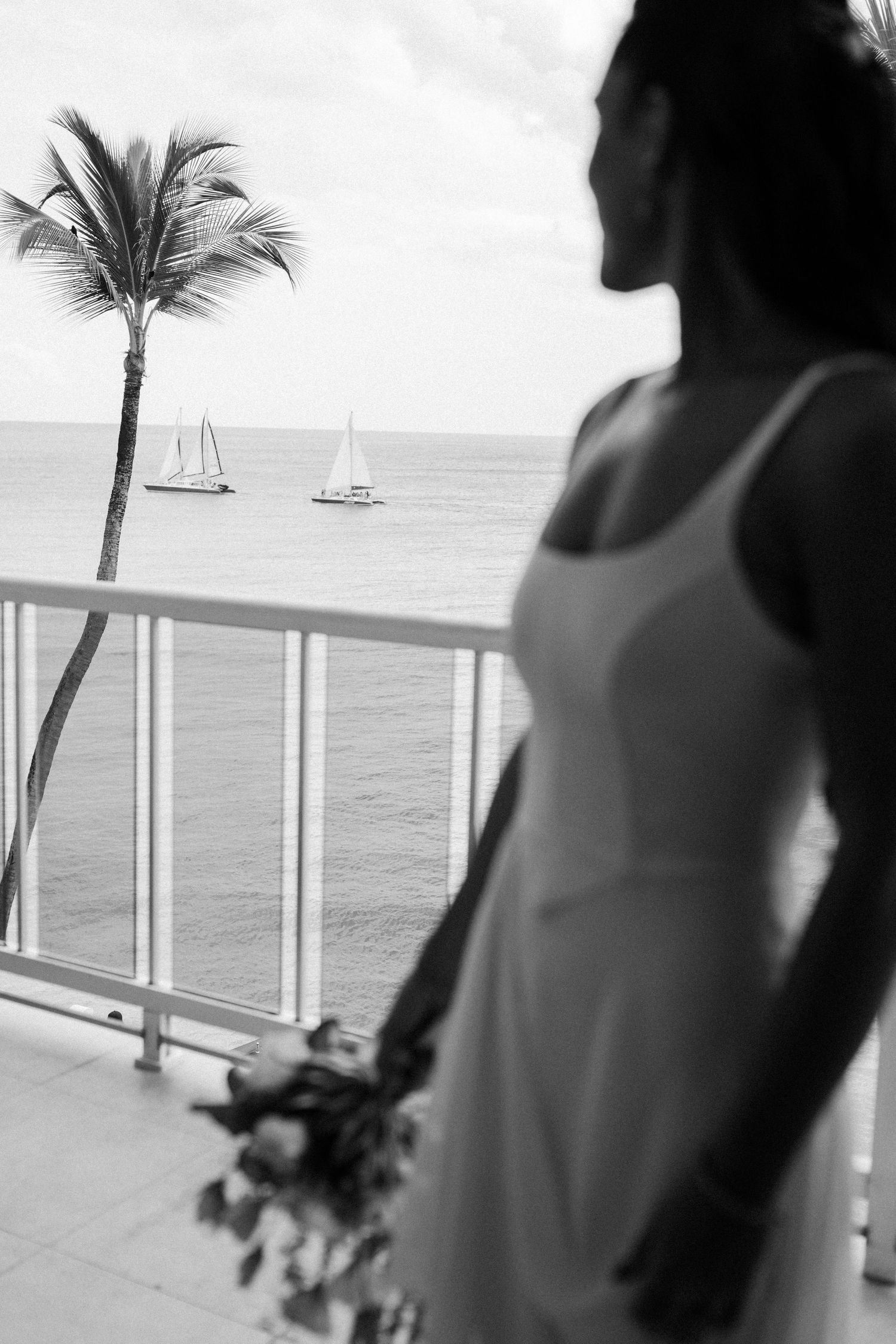 Bride in white gown holds bouquet on balcony overlooking ocean with sailboats and palm tree. Barbados Destination Wedding Photographer