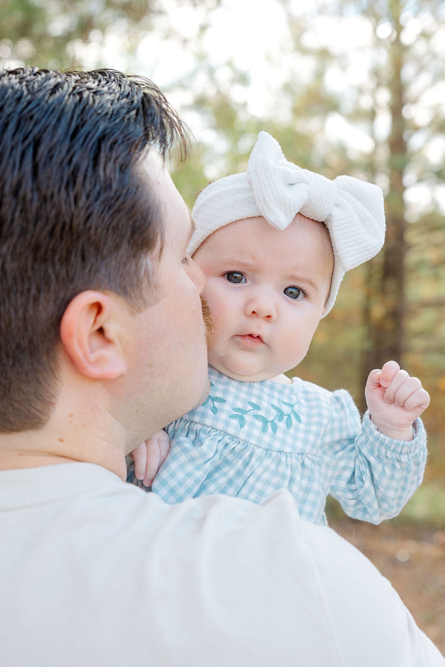 Father kissing baby girl with white bow headband in sunlit forest, tender family moment.  | Karina Thomson Photography Raleigh NC