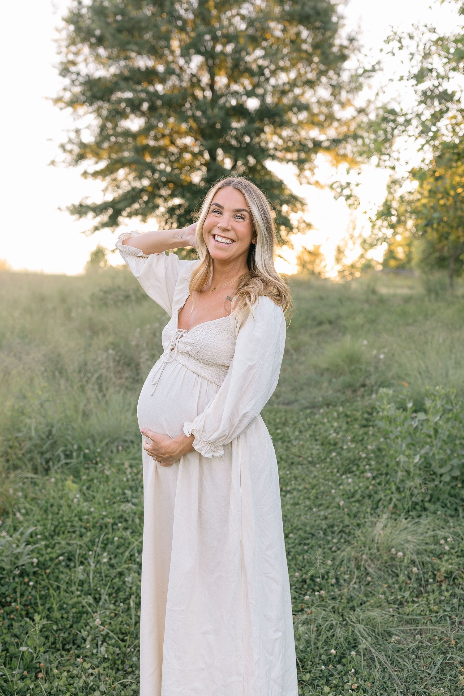 Smiling pregnant woman in white maxi dress holding baby bump outdoors at golden hour in a lush green field.