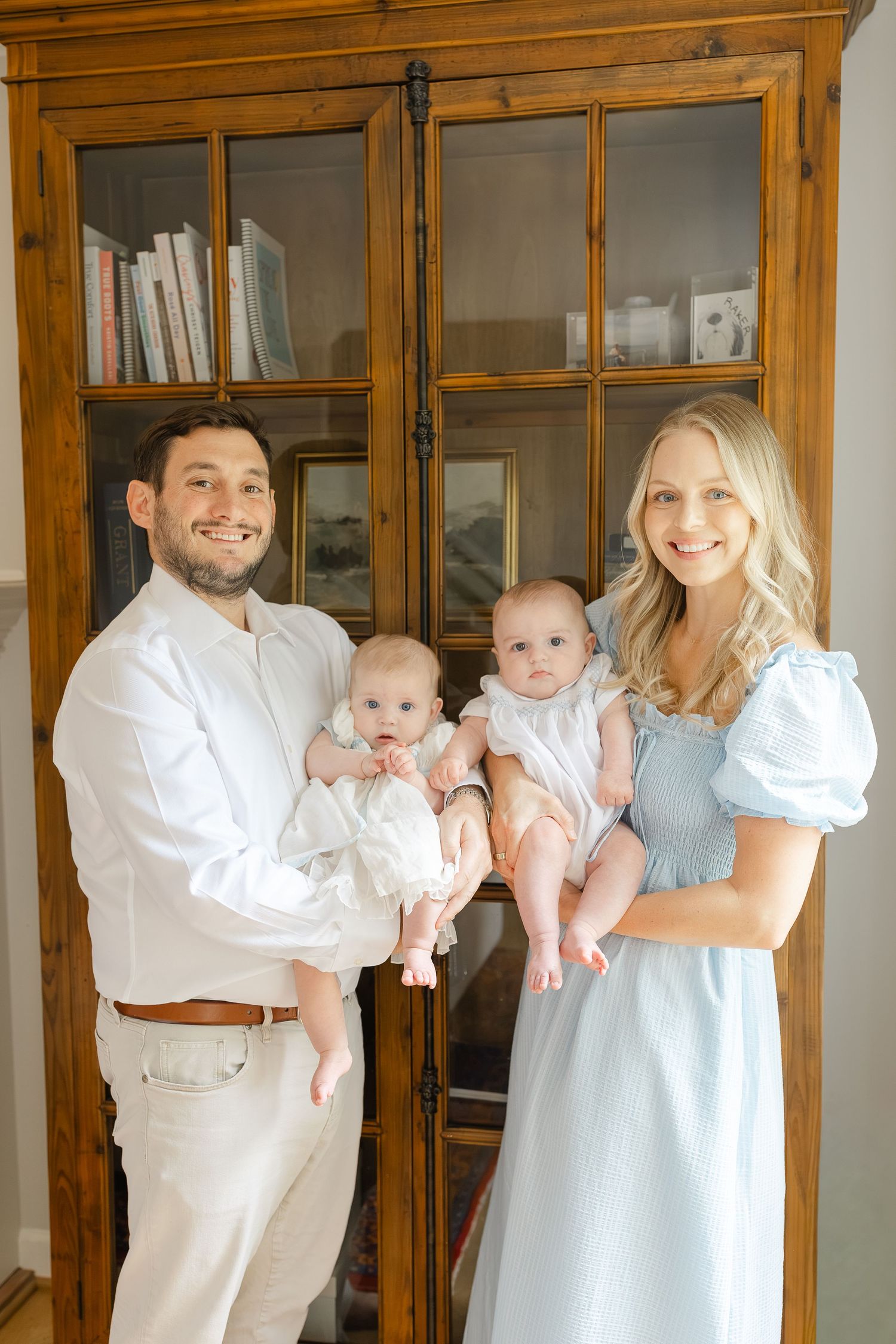 A smiling couple in light-colored outfits holds two babies in front of a wooden glass-door bookcase.