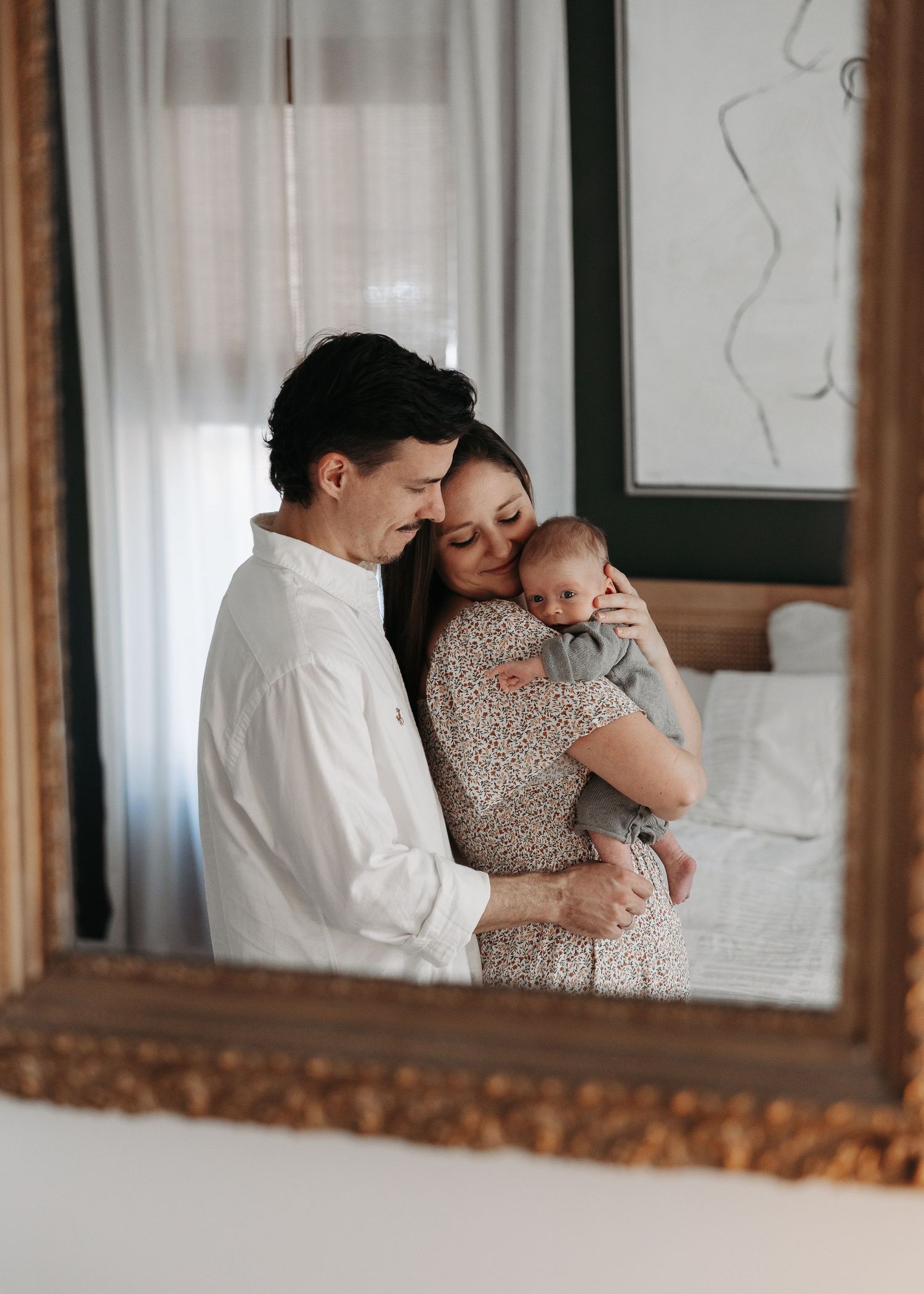 New parents tenderly embracing their newborn baby, reflected in an ornate gold-framed mirror.  | Karina Thomson Photography Raleigh NC