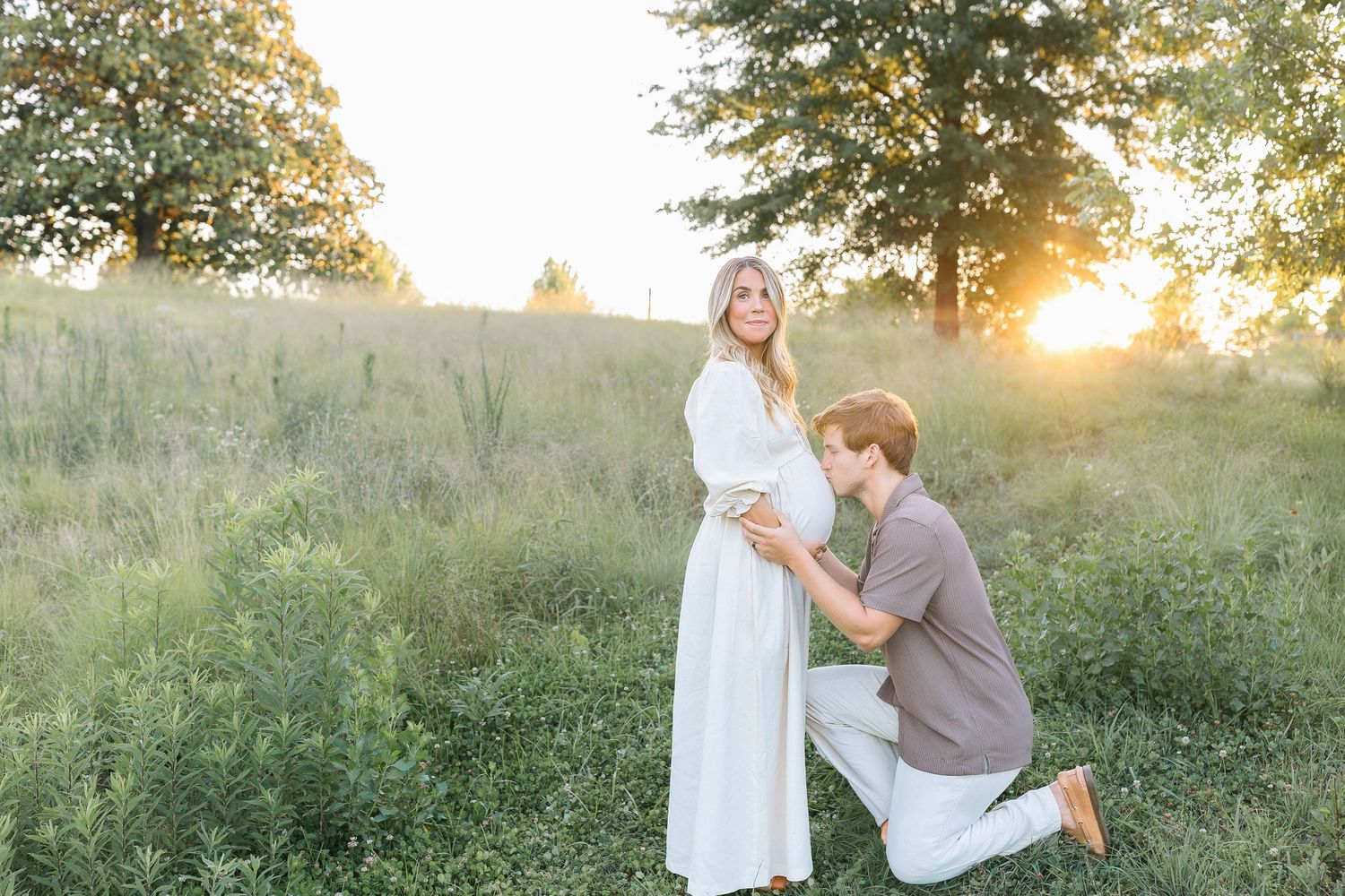 Couple in golden hour field, man kneeling kissing pregnant woman's belly in white dress.