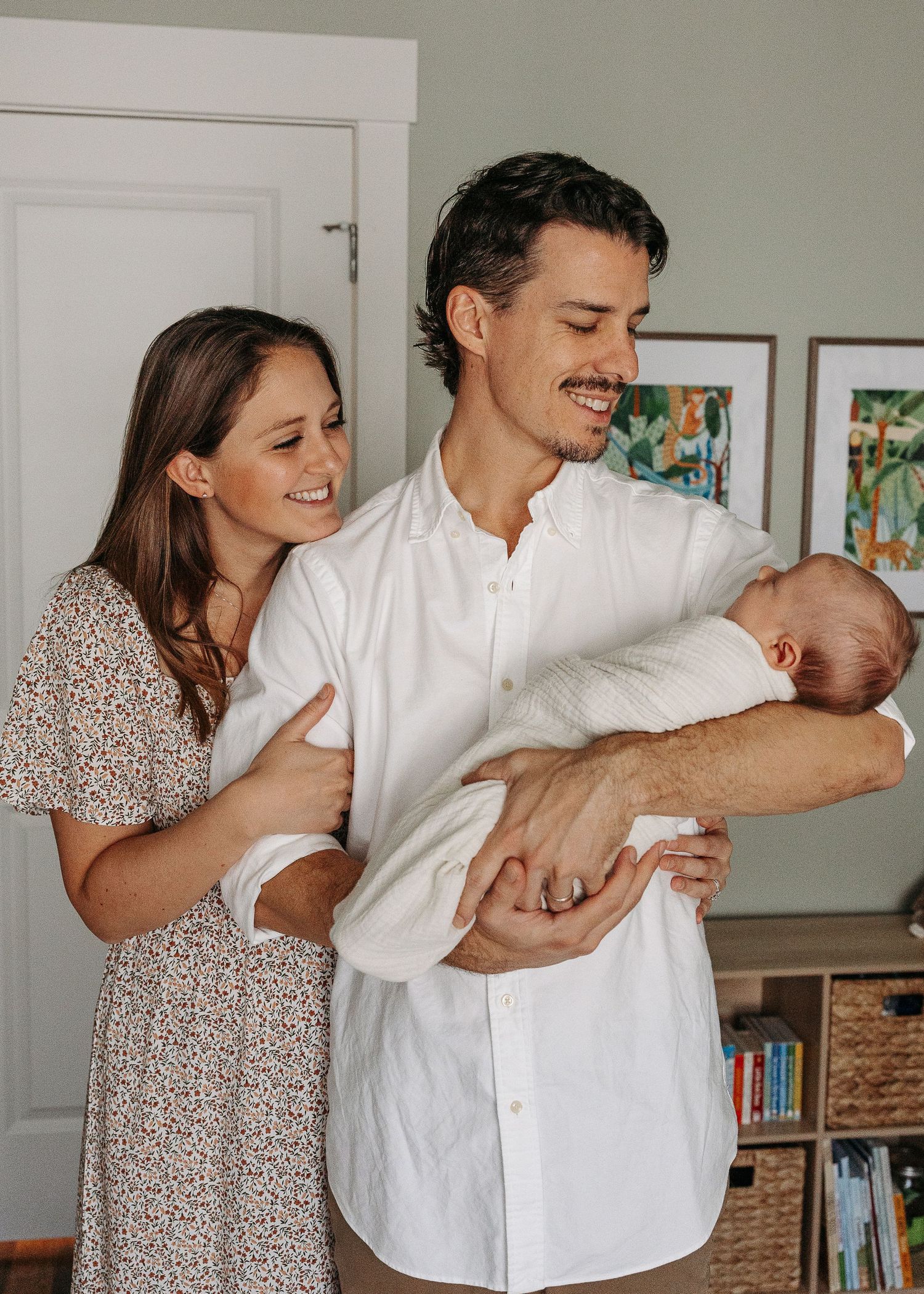 Young couple smiling together as the father holds their swaddled newborn baby at home.  | Karina Thomson Photography Raleigh NC