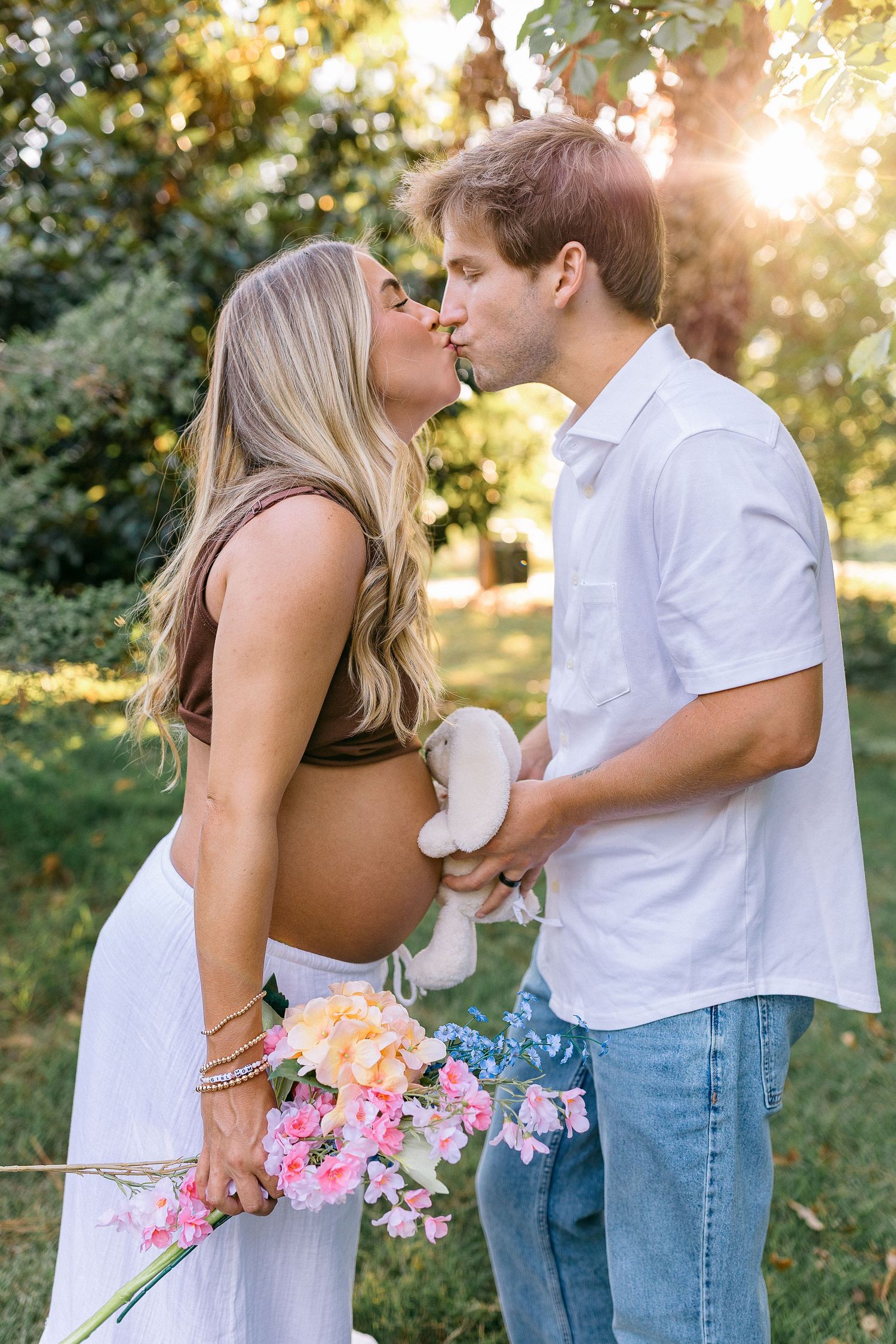 Pregnant couple kissing outdoors in golden sunlight, holding baby shoes and pink flowers, maternity photo session.