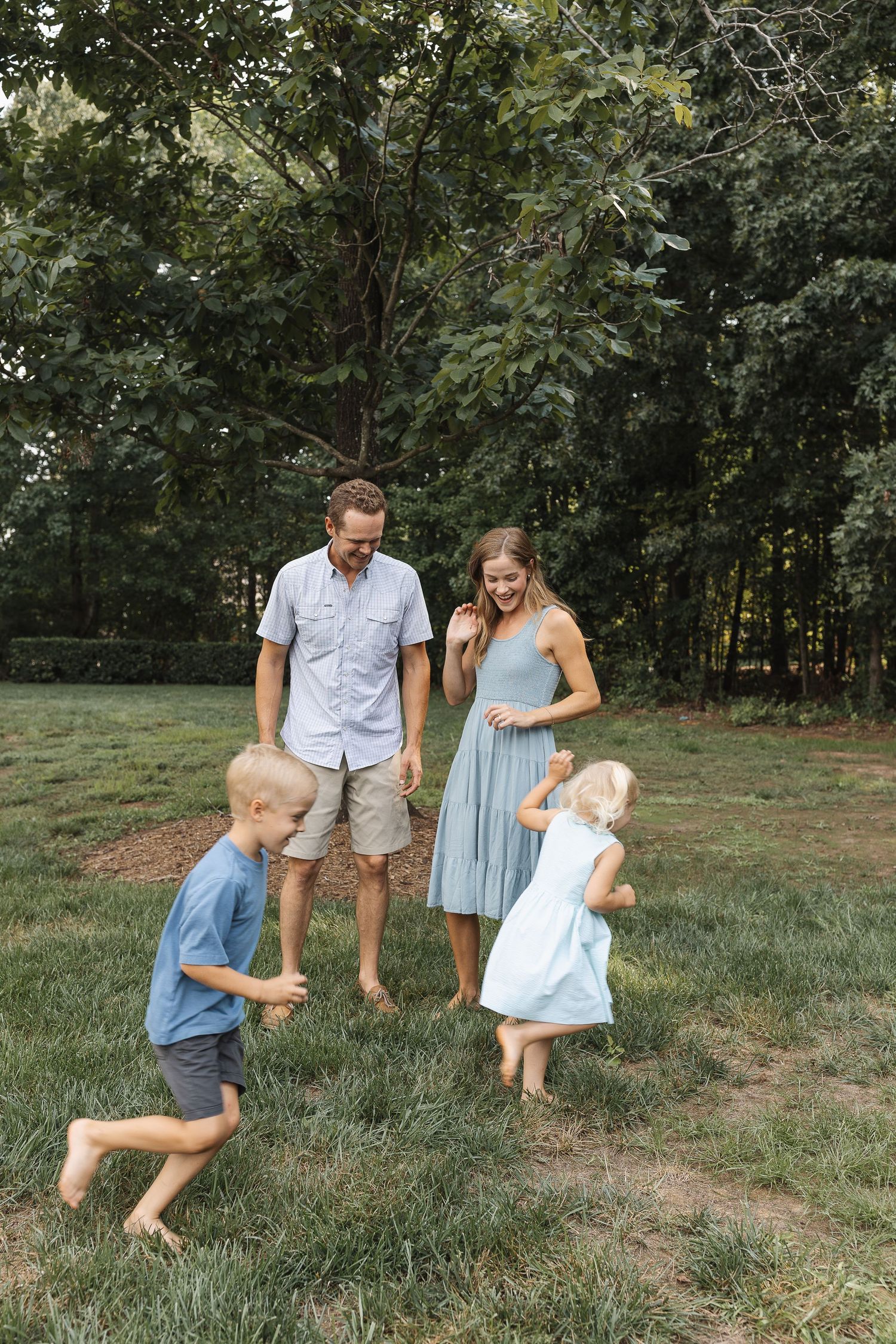 A happy family of four plays barefoot on green grass outdoors, parents smiling as two young children run around them.
