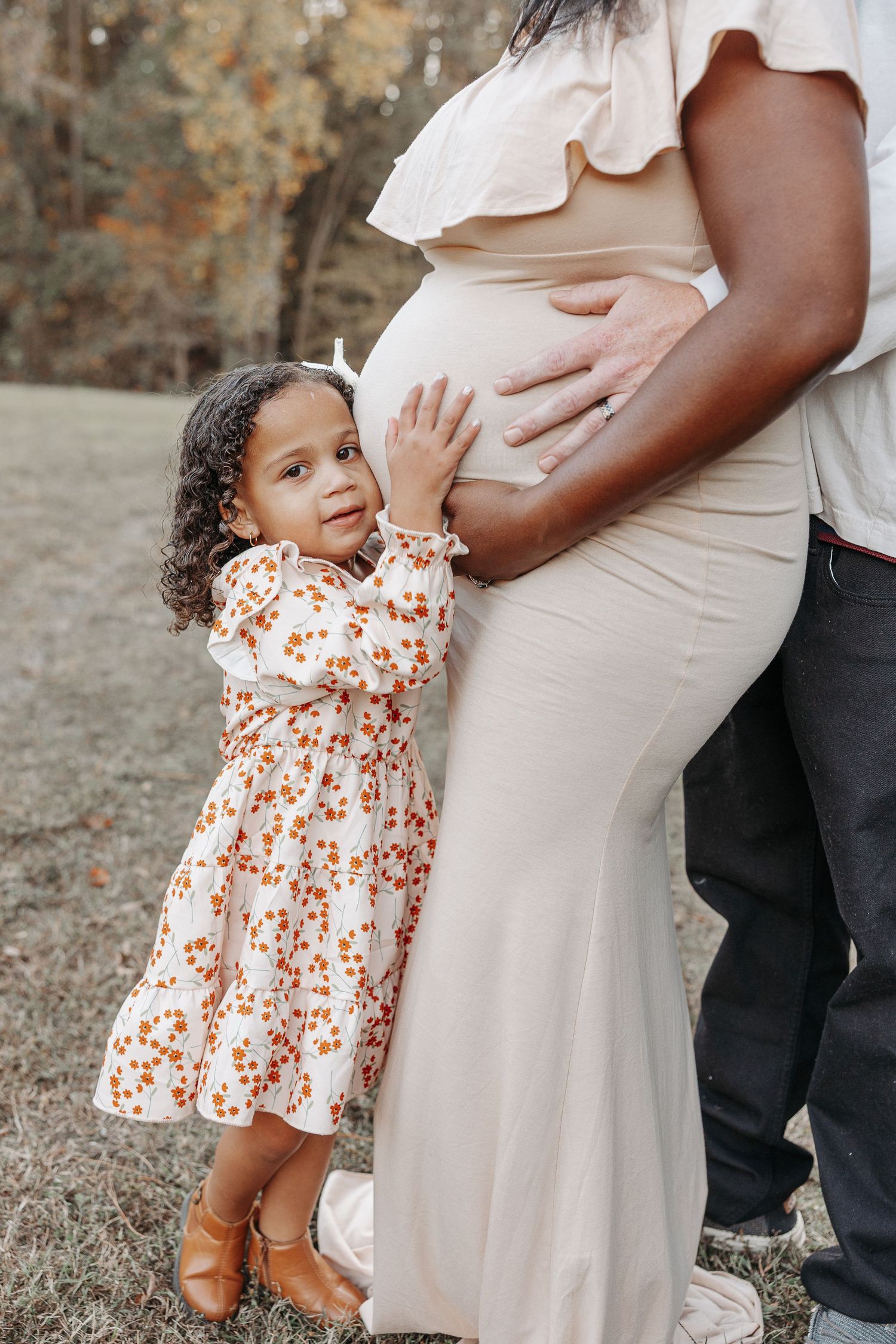 Young girl in floral dress touches pregnant mothers belly outdoors in autumn setting.  | Karina Thomson Photography Raleigh NC