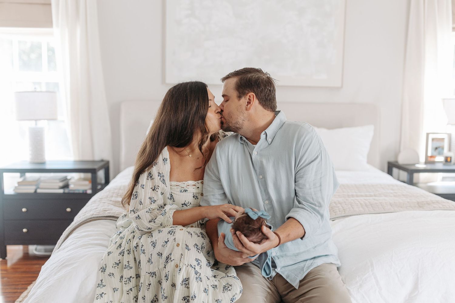 Couple shares a kiss on a bed while holding their newborn baby in a bright bedroom.  | Karina Thomson Photography Raleigh NC