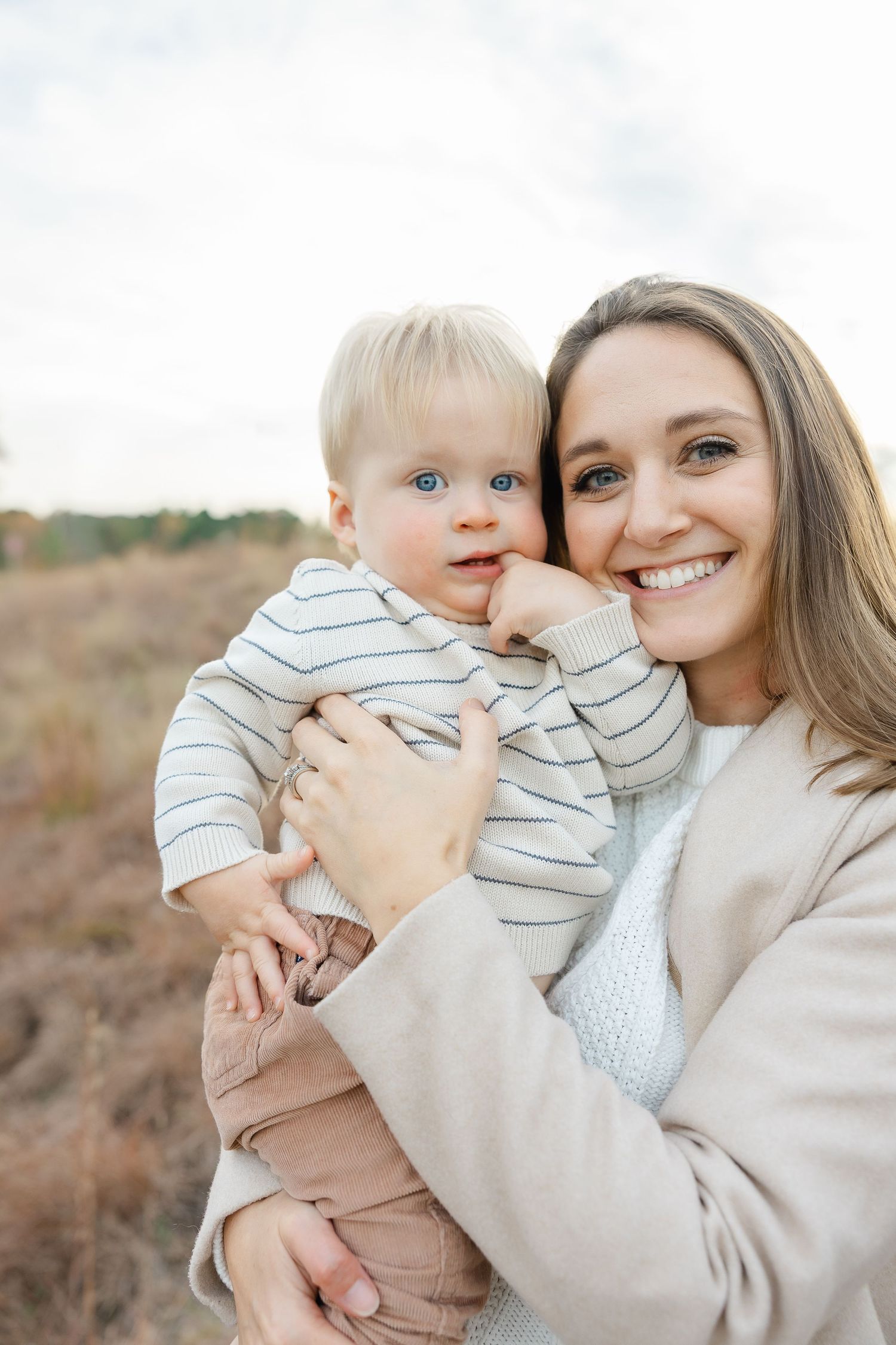 A smiling mother holds her wide-eyed baby outdoors in an autumn field setting.  | Karina Thomson Photography Raleigh NC