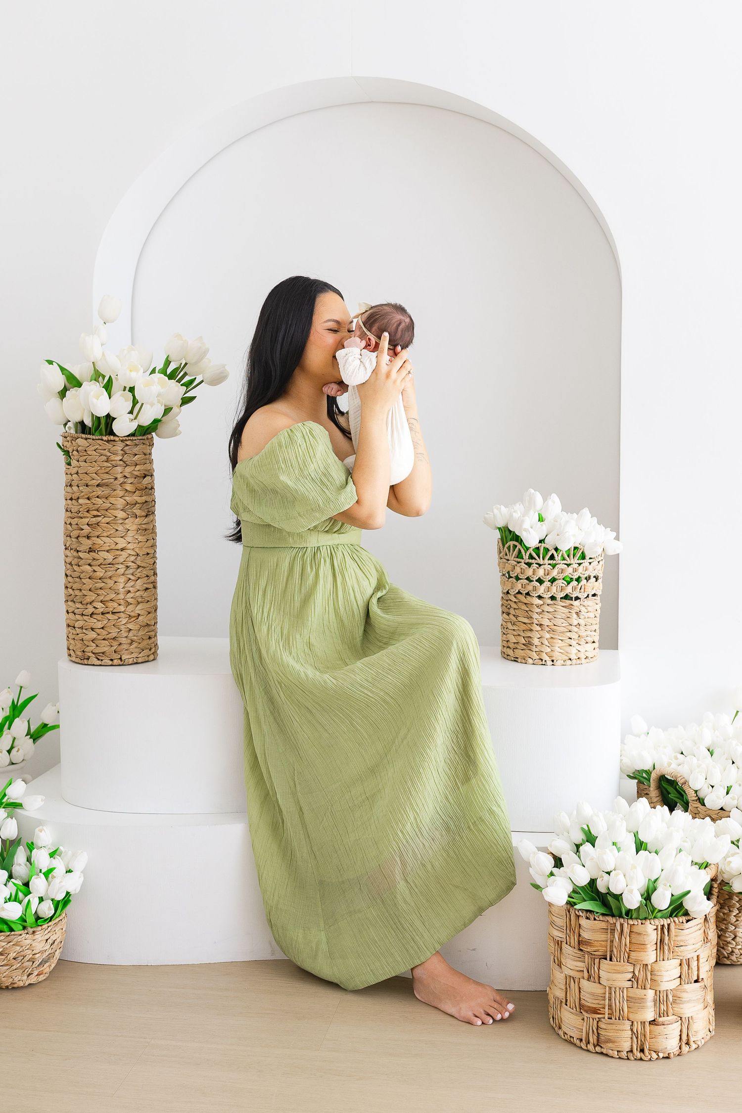 A woman in a green dress kisses her newborn baby surrounded by white tulips in wicker baskets.