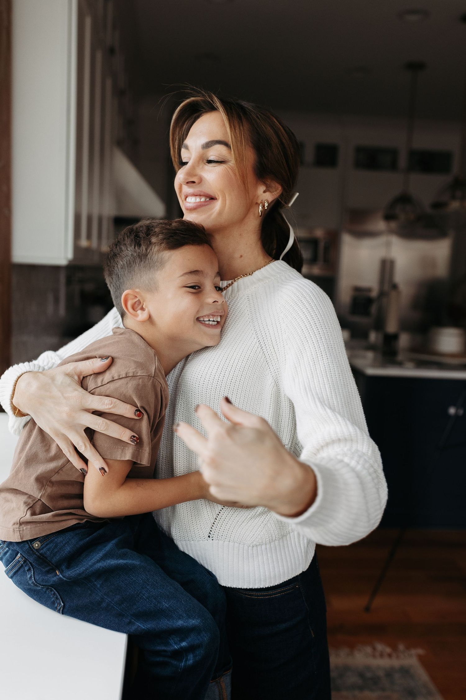 A smiling mother hugs her young son warmly in a cozy home kitchen setting.