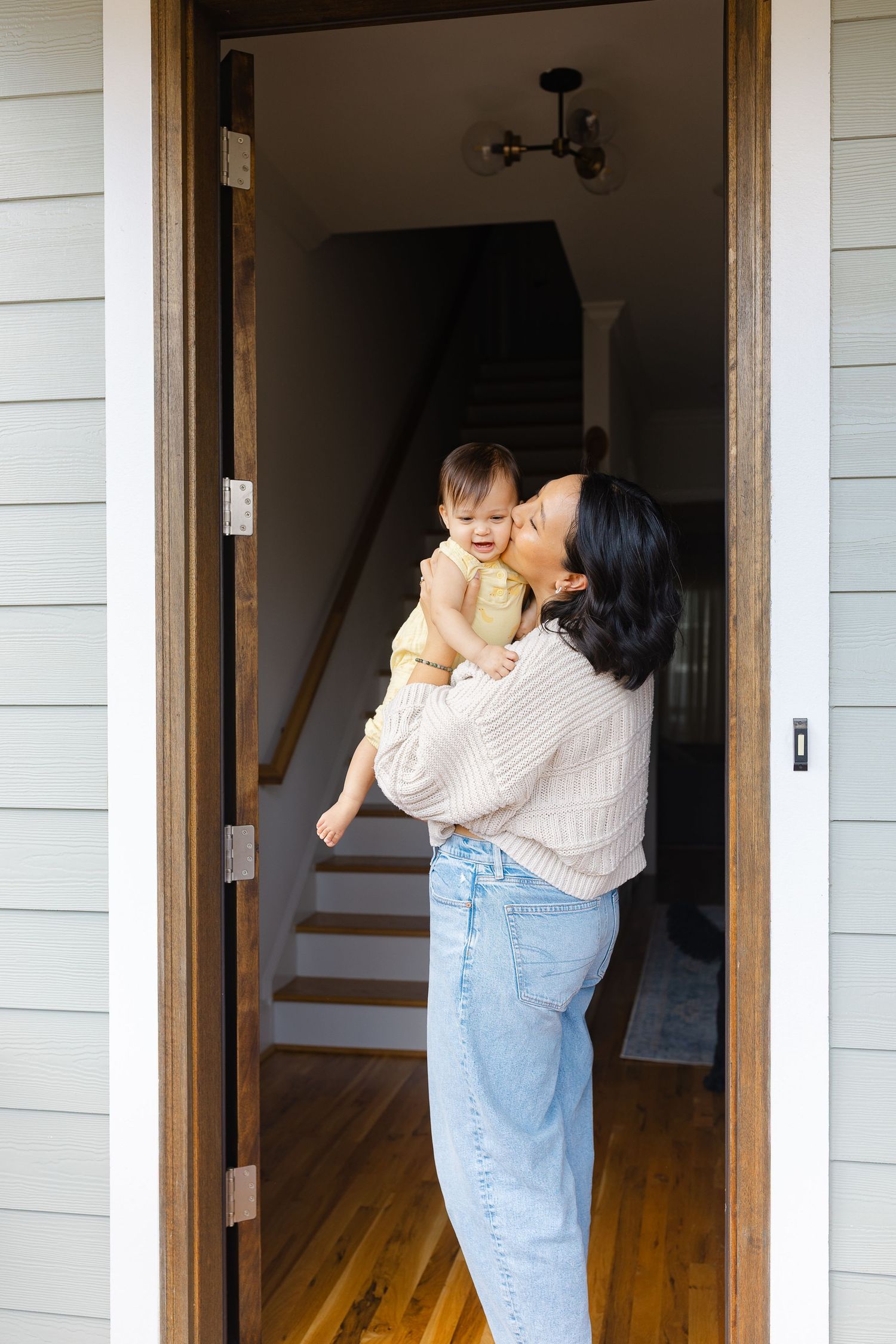 A mother kisses her smiling baby while standing in a charming home doorway.  | Karina Thomson Photography Raleigh NC