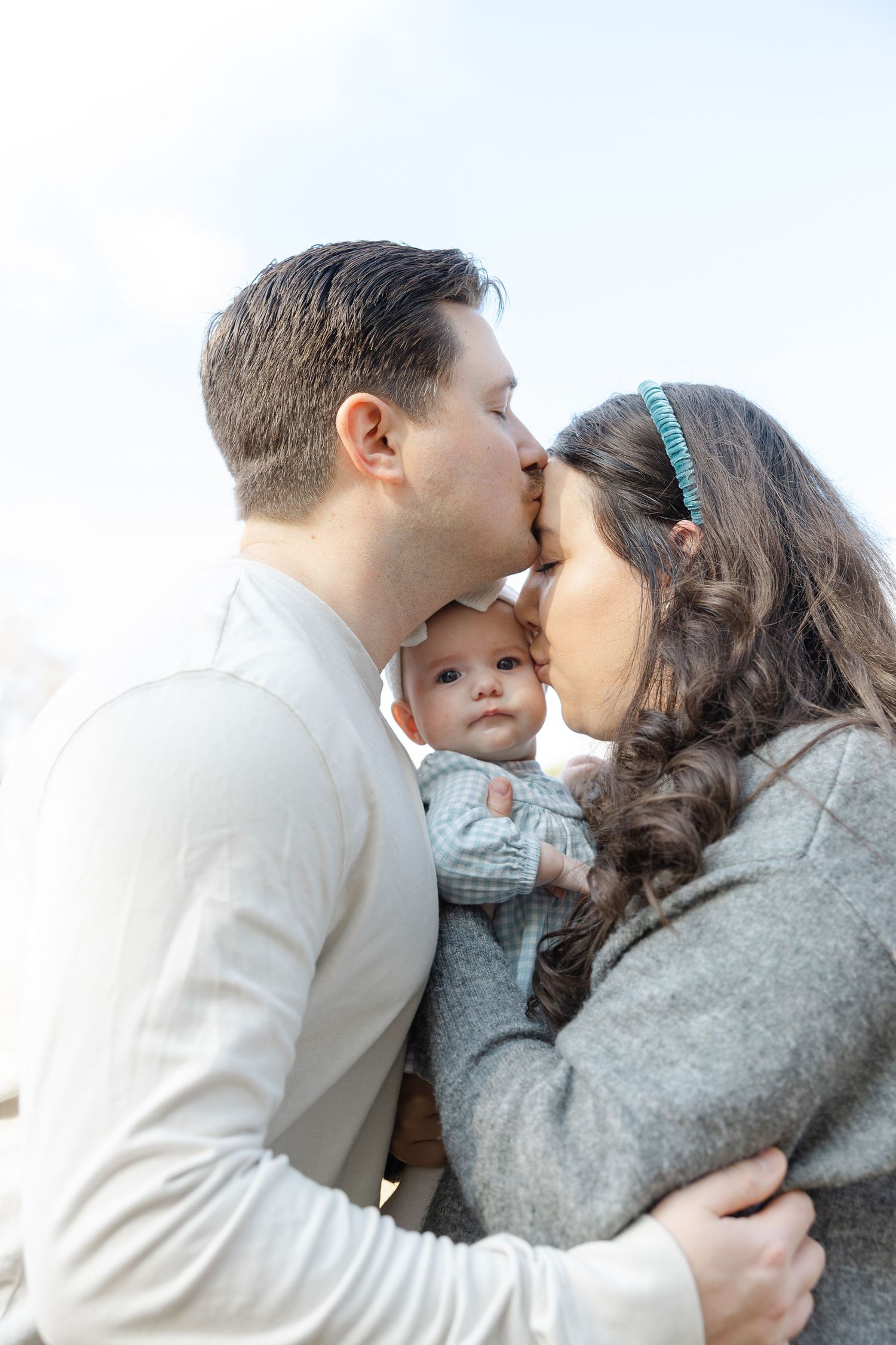 Couple sharing a kiss while holding their baby outdoors against a bright sky background.  | Karina Thomson Photography Raleigh NC