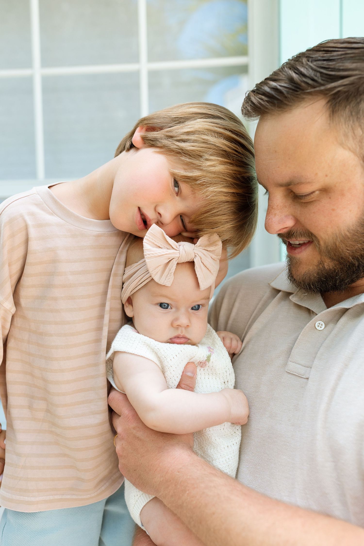 A bearded father holds a baby girl with a bow headband while an older blonde child leans in close, smiling warmly.