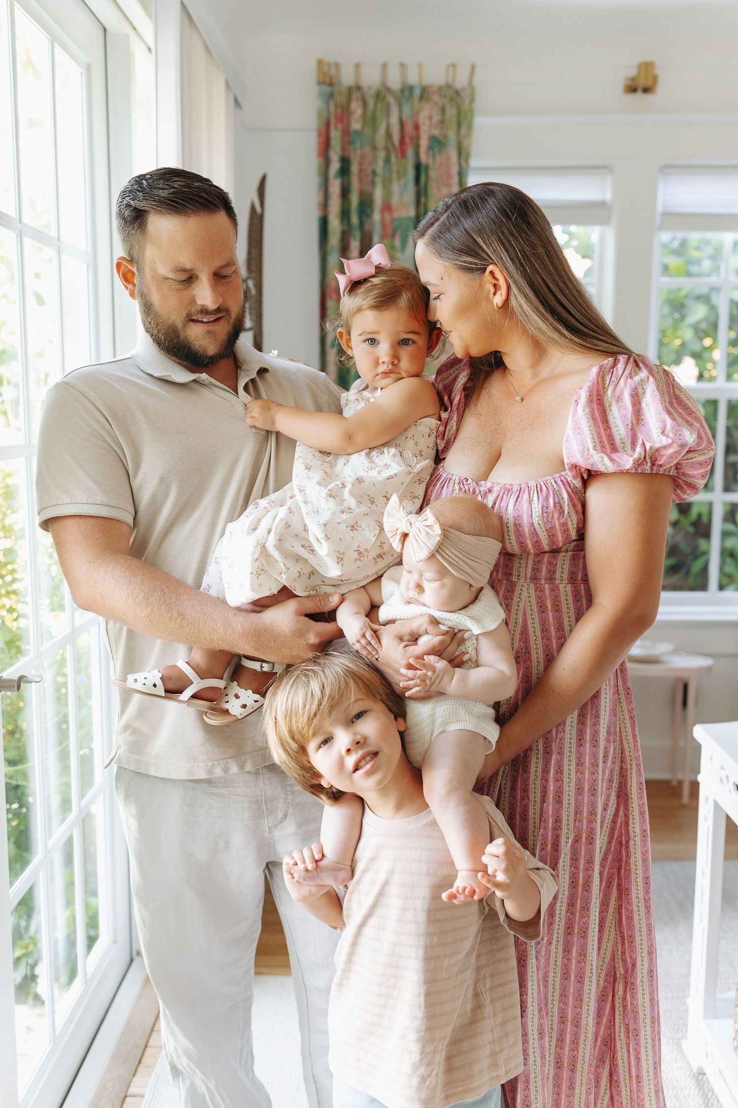 Happy family of five with two toddlers and a baby posing indoors by bright windows in a sunlit room.