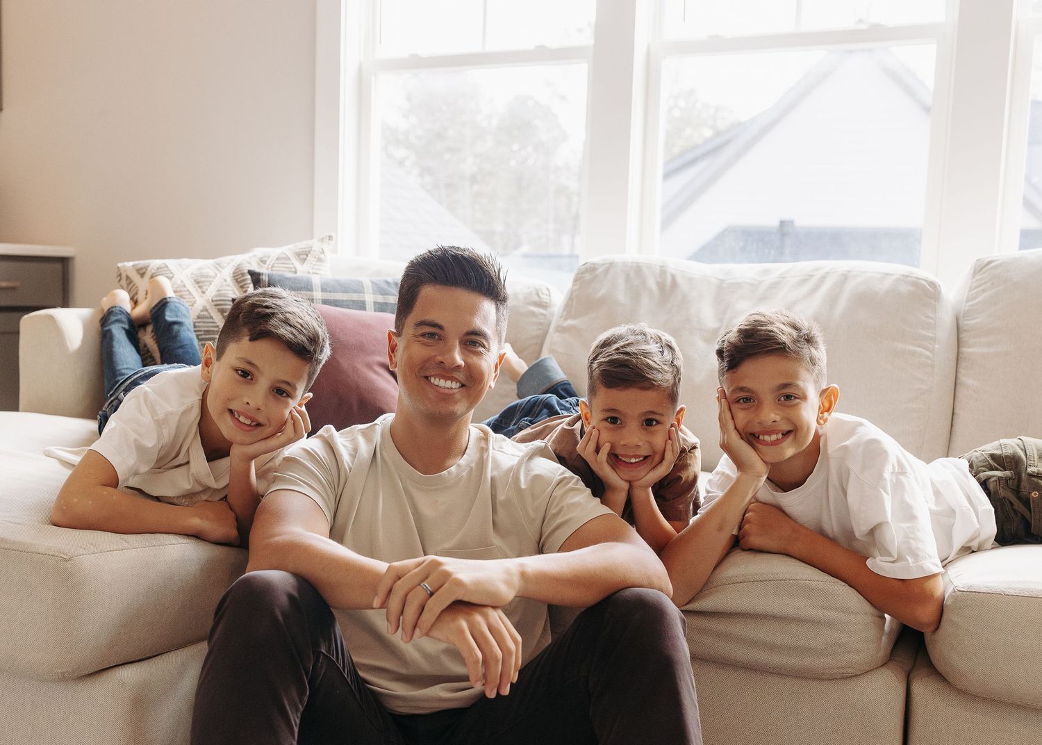 A father smiles with his three sons while relaxing on a couch in a bright room.  | Karina Thomson Photography Raleigh NC